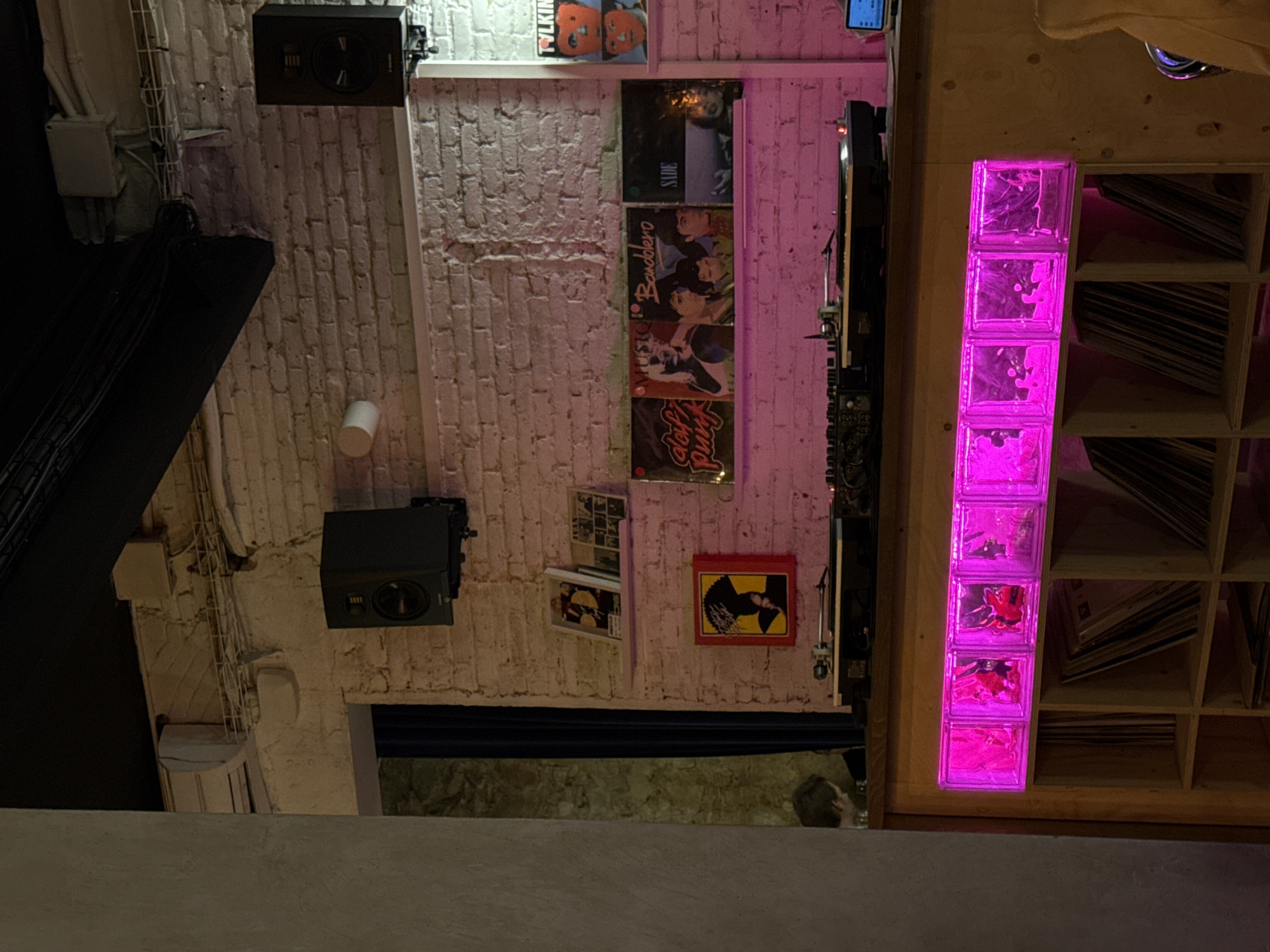 The image depicts an interior space in Barcelona, Spain, showcasing a wall adorned with framed vinyl records and a speaker system. The wall is constructed from white brick. Additional records are displayed on a shelf system with pink lighting. The space appears to be a record store or a music venue.