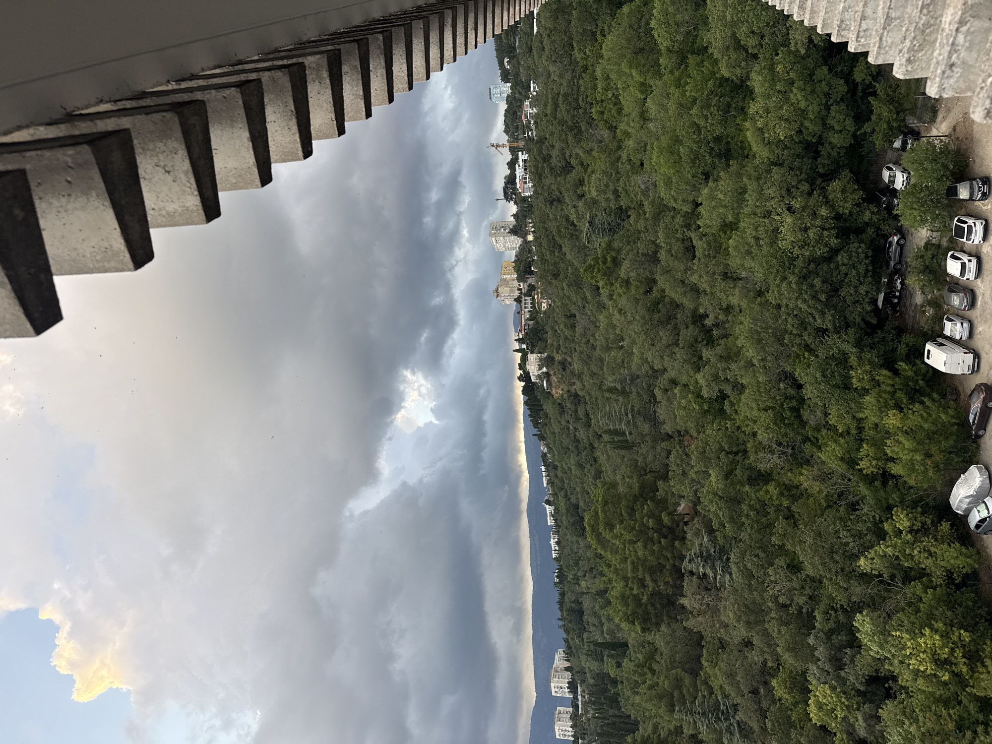 The image shows a view of a densely forested hillside with buildings on top, overlooking a cloudy sky. In the foreground, a concrete structure, possibly a balcony or a building facade, with a textured pattern of horizontal rectangular blocks, partially frames the scene from the right. The hillside is covered in lush green trees, with some taller cypress-like trees interspersed among the deciduous foliage. Several buildings, appearing to be residential or hotel complexes, are visible scattered on the upper slopes of the hill, some with white facades and others with lighter colors. The sky above is filled with dramatic, dark gray clouds, with a sliver of lighter, possibly sunlit, sky visible towards the left edge. The overall impression is a somewhat overcast day with a view of nature and urban development on a hillside. A small parking area with several cars is visible at the bottom of the hill. The provided context suggests the location is the city of Yalta, Ukraine.