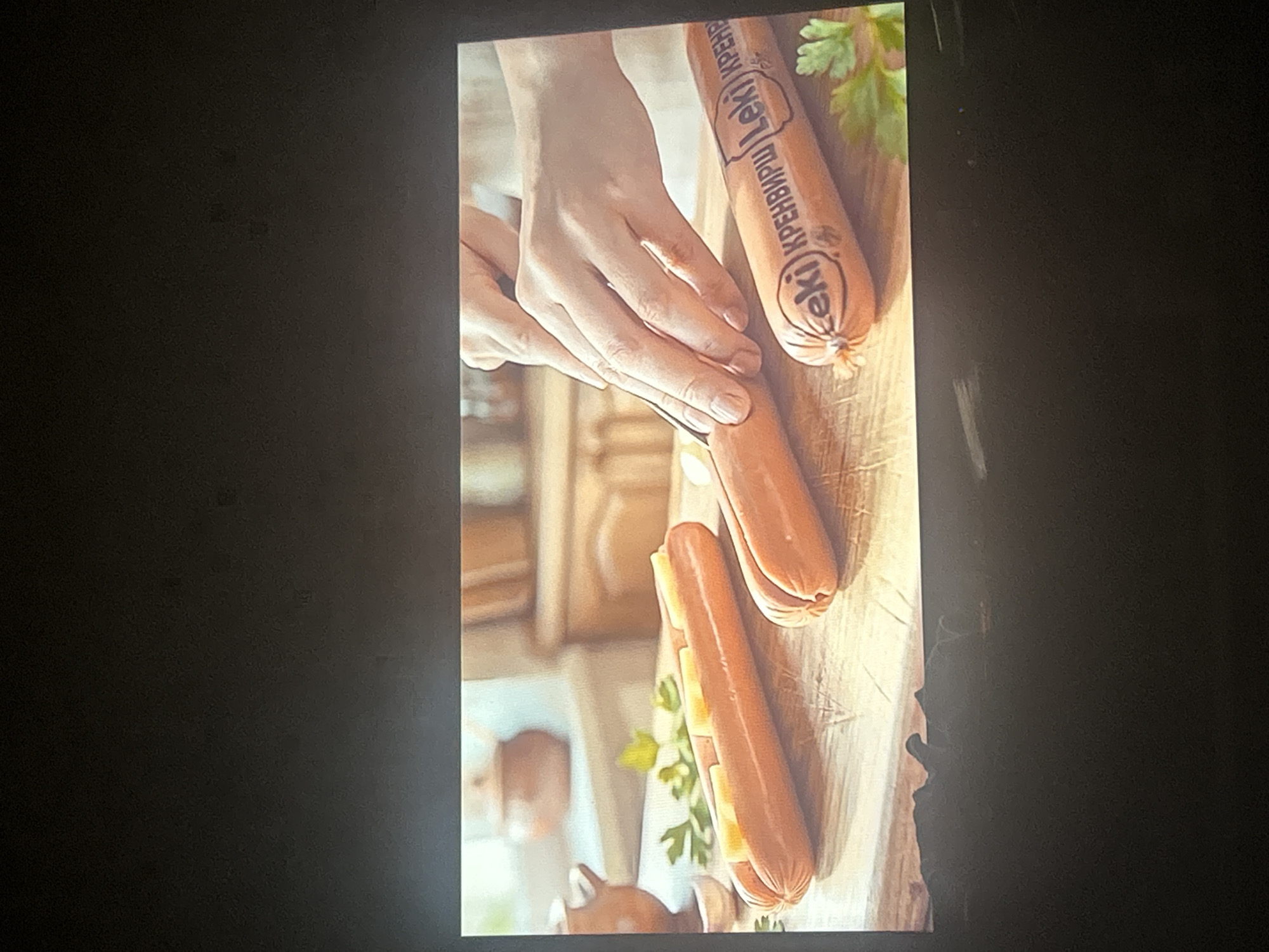 The image shows three sausages on a wooden surface, with a pair of hands preparing to slice one of them. The sausages are orange in color and appear to be of the hot dog variety. Two of the sausages are whole, while the third one is partially sliced open and filled with pieces of cheese. There are also sprigs of parsley and a small bulb of garlic visible on the wooden surface. In the background, there are kitchen cabinets and pottery, suggesting a kitchen environment. The overall scene depicts food preparation. The image is a still frame from a video, and the location in Sofia, Bulgaria is not discernible from the visual information provided.