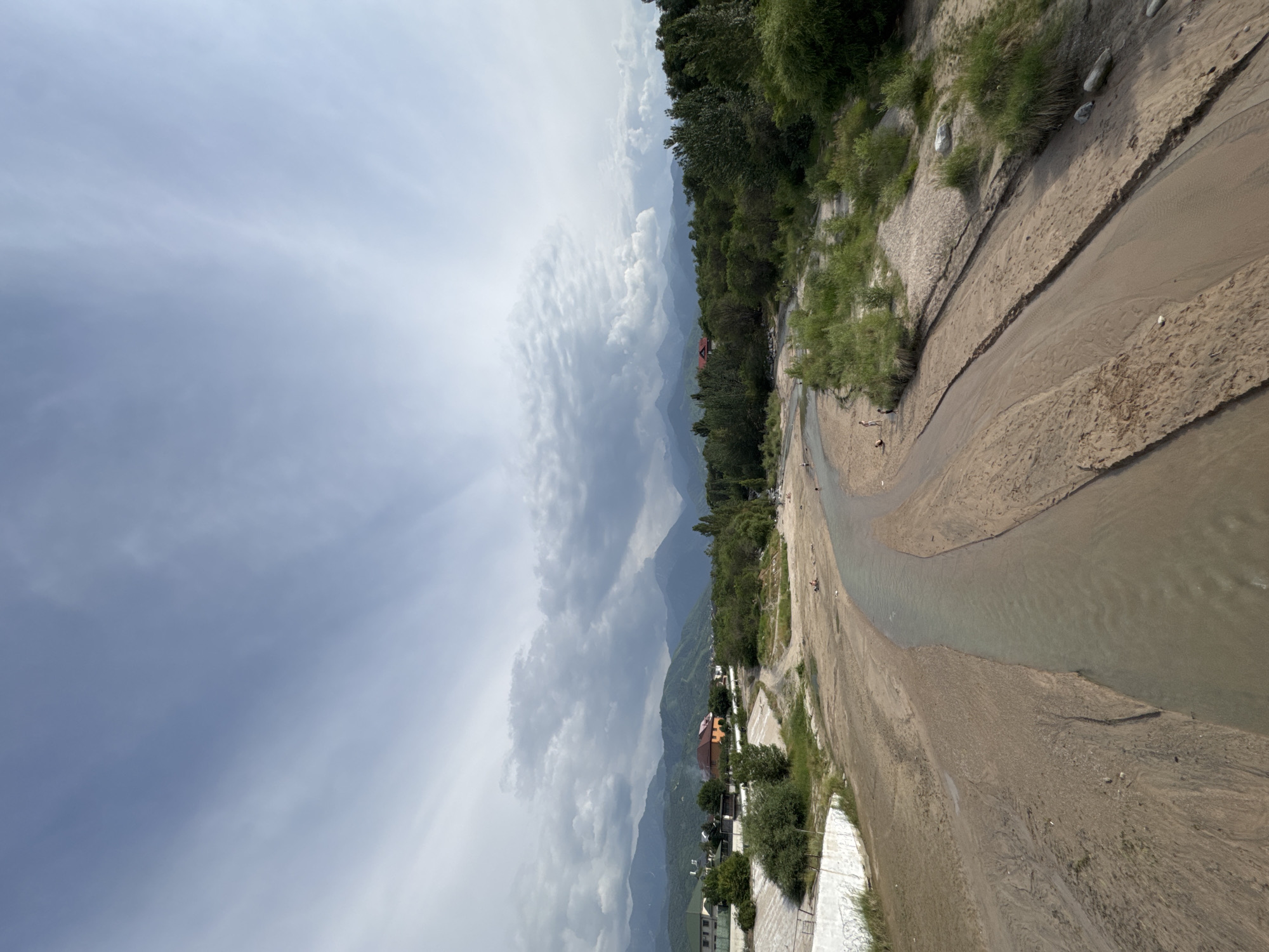 The image shows a wide, shallow riverbed with a narrow stream of water flowing through it. The riverbed is composed of sand and gravel, with some vegetation growing along the banks. In the background, there are mountains covered in greenery, and several houses with red roofs are visible on the slopes. The sky is cloudy, with rays of sunlight breaking through. The scene appears to be a natural landscape within a residential area. Based on the scenery, including the distinctive mountains and the architecture of the houses, this location is likely the Malaya Almatinka River or a similar river in the foothills of the Trans-Ili Alatau mountains near Almaty, Kazakhstan.