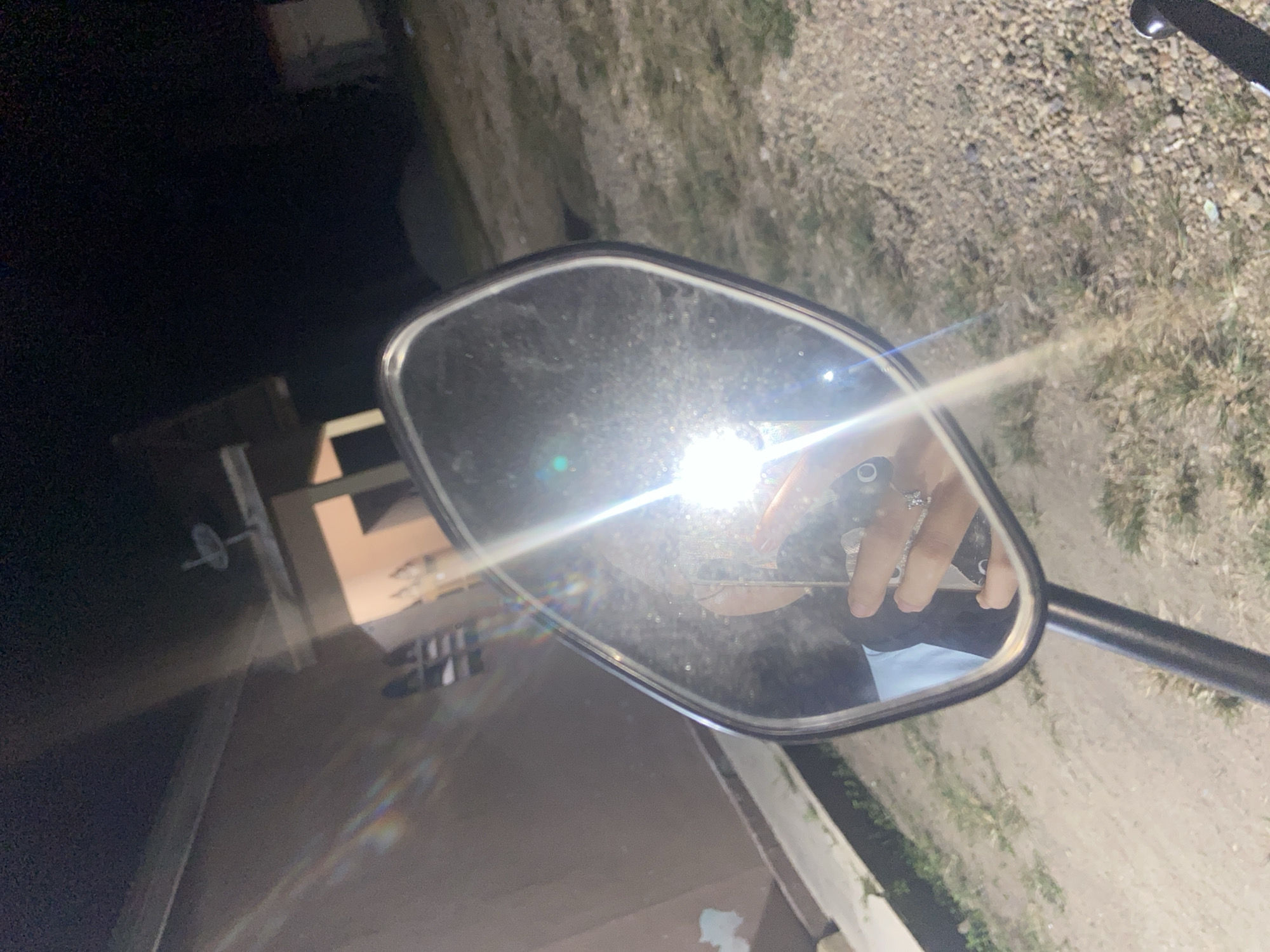 The image shows a motorcycle mirror reflecting a person's hand and arm, holding a smartphone. The person is wearing a blue and black shirt and multiple rings on their fingers. The mirror is dirty, with visible smudges and dust particles. The background is dark, suggesting it is nighttime. A bright light source, possibly a camera flash or streetlight, is reflecting strongly in the mirror, causing lens flare and obscuring details of the surroundings. The environment appears to be an outdoor area with gravel and some sparse vegetation. Based on the provided information, the location is the city of Sekar Mawar, Indonesia.