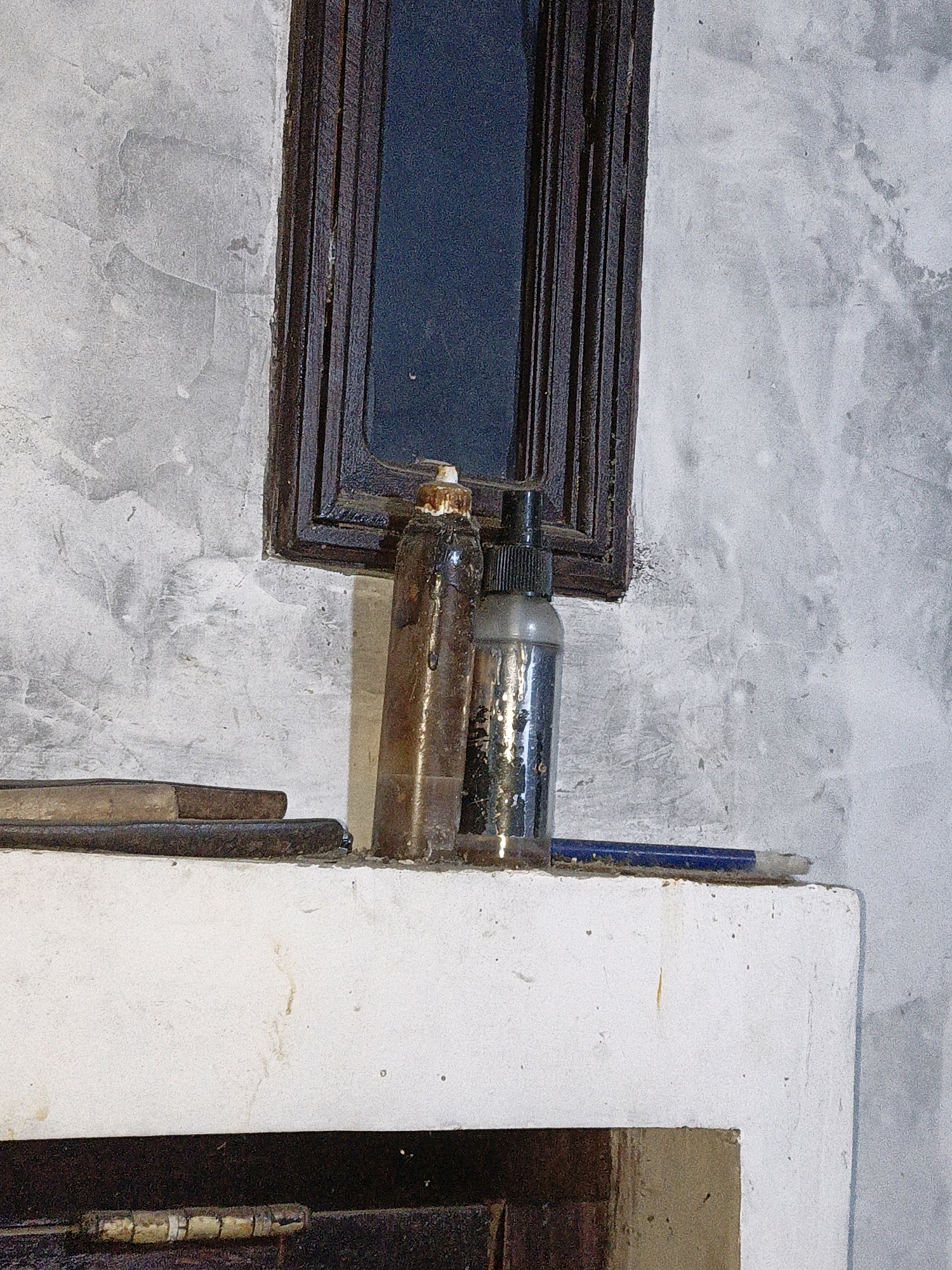 The image depicts an indoor scene with two bottles on a white ledge. The bottle on the left is tall and brown, with a cap that appears to be made of melted wax or sealant. The bottle on the right is shorter, clear, and contains a silver liquid, with a black cap. Both bottles show signs of wear and tear, with drips and smudges on their surfaces.
To the left of the bottles, two wooden objects are stacked horizontally on the ledge. These appear to be tools or implements, with one being darker and more worn than the other. A blue pencil lies on the ledge in front of the bottles, pointing to the right.
The background consists of a textured wall, with a dark rectangular frame in the upper center of the image. This frame has a dark, glassy surface behind it, suggesting a window or a similar opening. The wall itself has a rough, uneven texture, with patches of light and dark grey. The white ledge appears to be part of a structure, possibly a fireplace or a shelf, and there is a dark opening beneath it.
The overall scene suggests an area where tools or art supplies might be stored, possibly in a workshop or a craft space. The lighting is somewhat dim, highlighting the textures and details of the objects. No people are visible in the image. The location is described as Cikal, Indonesia.