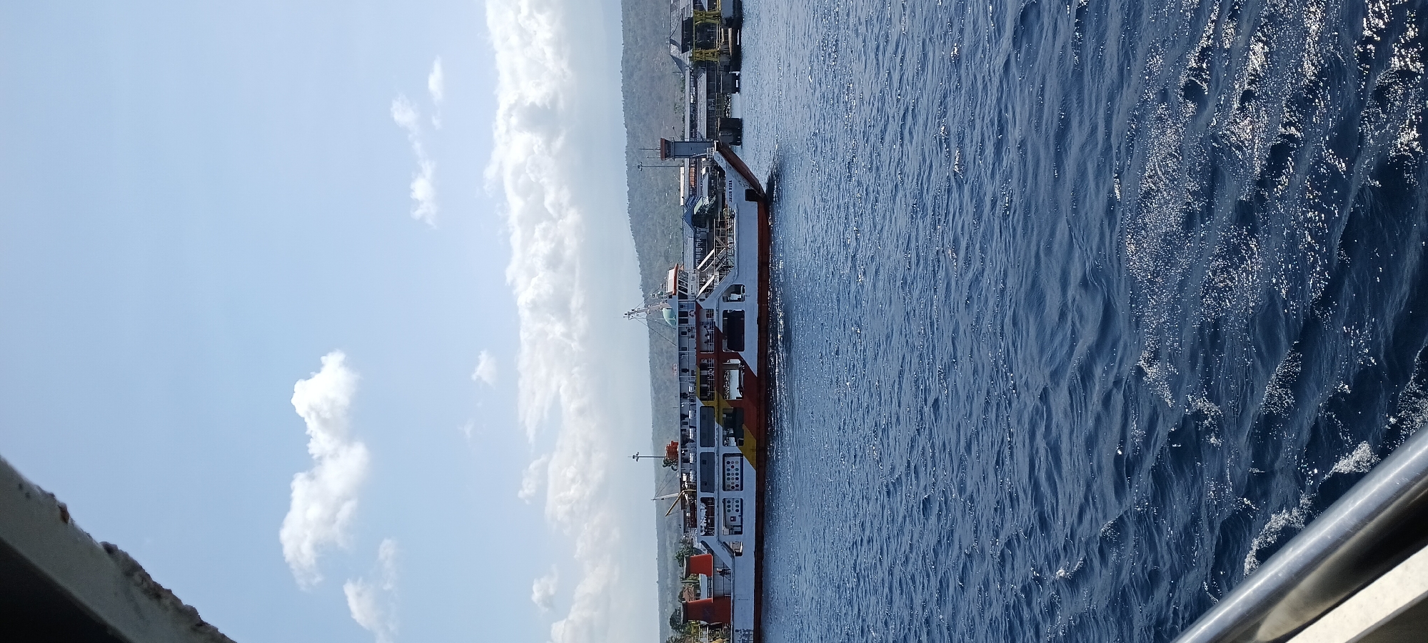 A ferry boat is docked at the Port of Ketapang, Banyuwangi, Indonesia. The ferry is white with red and yellow accents. It is a sunny day with a clear blue sky and some white clouds. The water is blue and choppy. In the background, there are hills and trees, and the port facilities are visible.