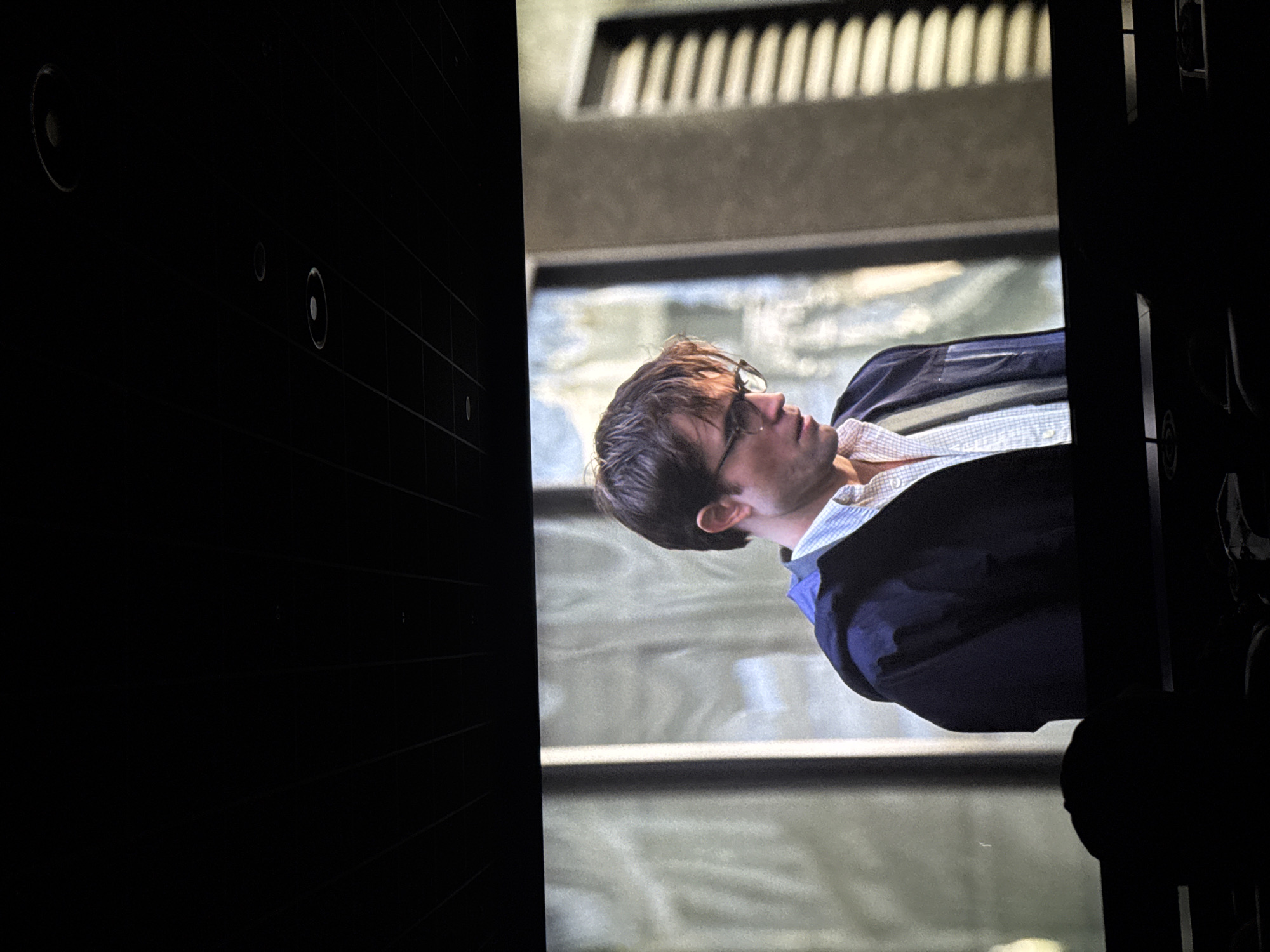 The image shows a screen displaying a man wearing glasses, a light-colored shirt, and a navy jacket. The background behind the man appears to be an urban environment, possibly with reflections of water. The surrounding area is dark, revealing features such as wall panels with circular recesses on the left side and a dark structure on the right side, which seems to be part of the room housing the screen. There is no information to determine the location in the city of Warsaw, Poland.