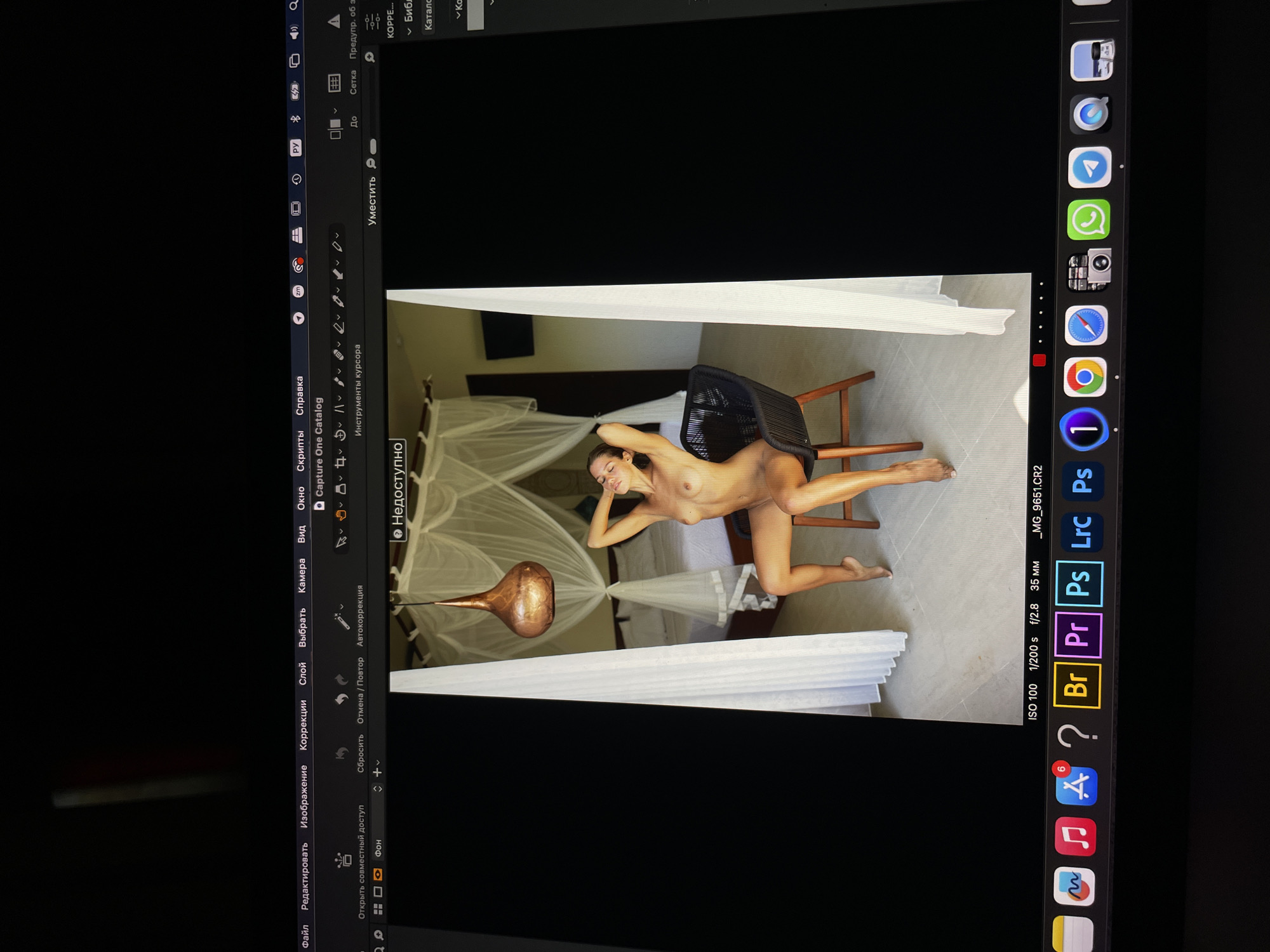 Here is a matter-of-fact description of the scene:
The image is a photo of a computer screen, oriented vertically. The screen displays an image editing program interface, with an image of a nude woman visible in the center of the screen.
The woman is lying on a black chair in a room with a bed draped with white mosquito netting. A decorative lamp hangs from the ceiling. The room appears to be a hotel room.
The computer interface surrounding the image shows toolbars and menus in Russian, suggesting the location of the computer is in Russia.
No information or evidence specifically places this scene in the city of Kazan.