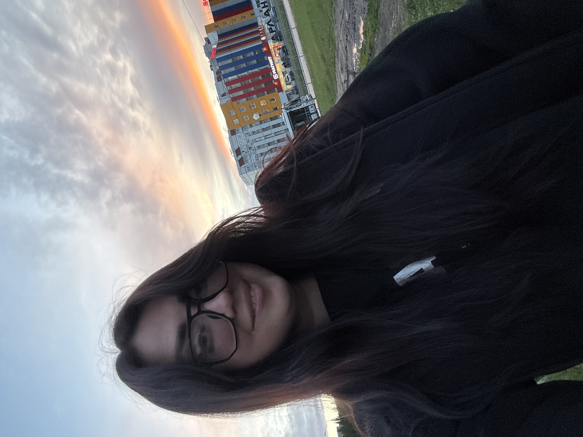 A young woman with long, dark hair and glasses is the focal point of this photograph, which is shot from a low angle, slightly off-kilter. Her face is partially visible, with a smile showing. She's wearing a dark-colored garment, possibly a jacket or sweater.
In the background, a dramatic sky featuring clouds tinged with shades of orange and pink suggests either sunrise or sunset. A tall, multi-colored building can be seen, with segments of white, yellow, red, and blue. Letters in what seems to be the Russian alphabet are visible on the building. A green area, likely grass or foliage, is also present in the background. The location is in a city of Unknown, Unknown.
