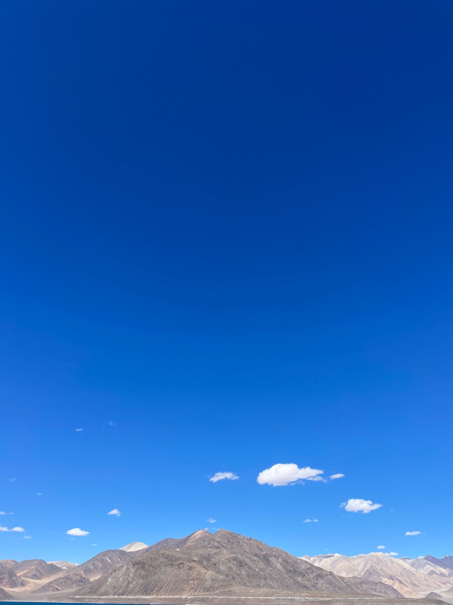 A wide-angle landscape photograph captures a vast, clear blue sky dominating the upper two-thirds of the frame. Below the sky, a range of rugged, arid mountains stretches across the horizon. The mountains display varied textures and colors, with shades of brown, tan, and grey, indicating a dry, rocky terrain. Patches of snow or lighter-colored rock are visible on some of the higher peaks. In the lower portion of the image, a sliver of a body of water, appearing as a deep blue, is visible to the left, suggesting a lake or large river. A few scattered, wispy white clouds are present in the sky, providing minimal interruption to the otherwise clear expanse. The scene appears to be located in a remote, mountainous region, likely at a high altitude, with the distinct geographical features suggesting a location like the Himalayas. This description is consistent with the landscape of Man, India, known for its dramatic mountain scenery. There are no people or man-made structures visible in the image.