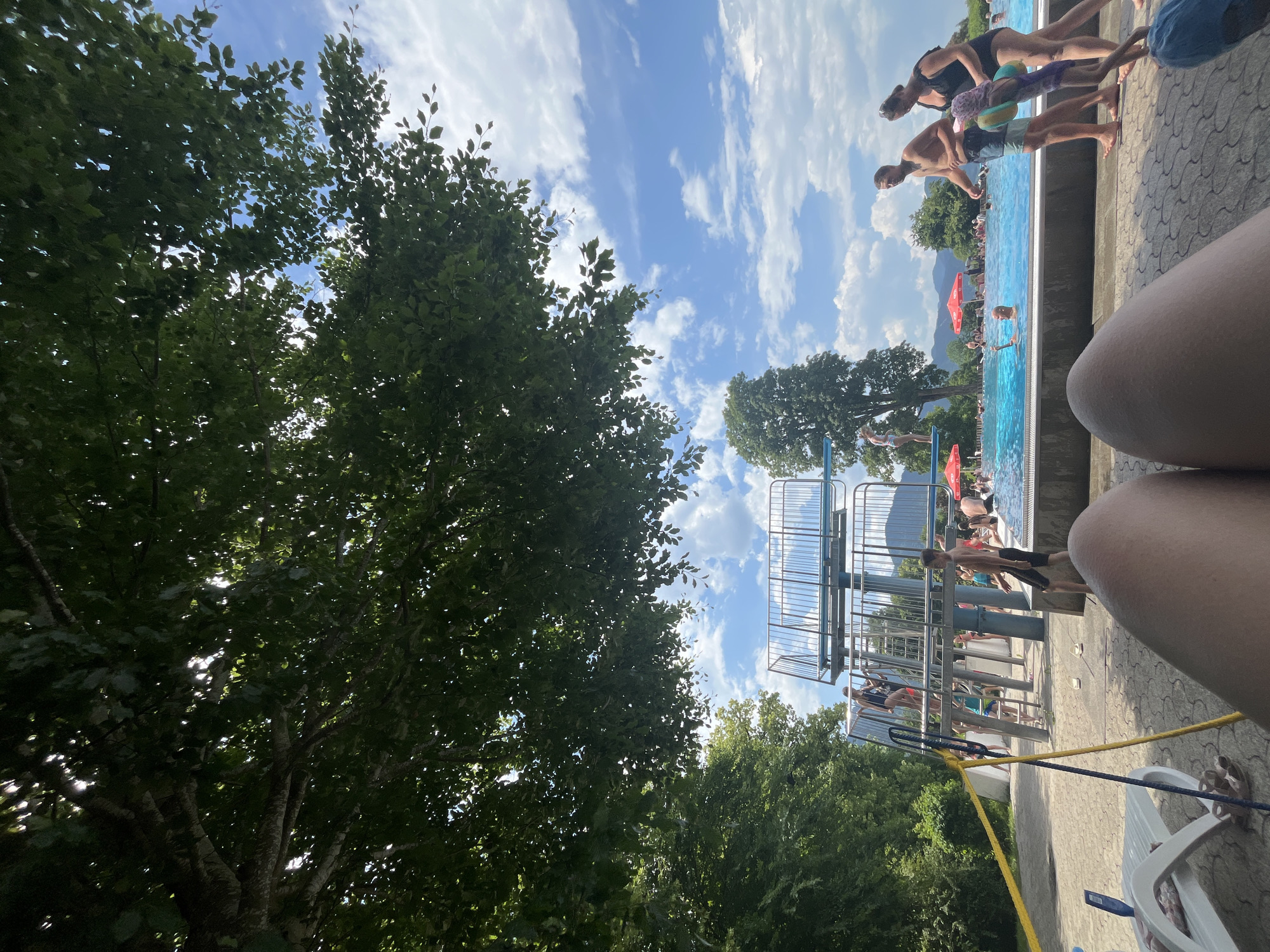 The image captures a scene at an outdoor swimming pool facility, likely in Oberammergau, Germany, given the presence of mountains in the background and the overall aesthetic. The perspective is from a low angle, as if someone is lying down on a lounge chair, with their legs partially visible in the foreground. The pool itself is a vibrant blue, surrounded by a paved area.
Several people are present around the pool. In the mid-ground, a diving tower with multiple platforms is visible, with a few individuals on its stairs and one person at the top, possibly preparing to dive. Children and adults are scattered throughout the pool area, some swimming, others standing by the edge. Two adults and a child are walking near the pool's edge, facing away from the camera.
To the left of the pool, there are trees and some lounge chairs, including the one partially seen in the foreground. The sky is a bright blue with scattered white clouds, and lush green trees frame the top of the image. Red umbrellas are also visible in the background, suggesting a recreational area. The overall impression is a sunny day at a public pool, with people enjoying the water and the warm weather.