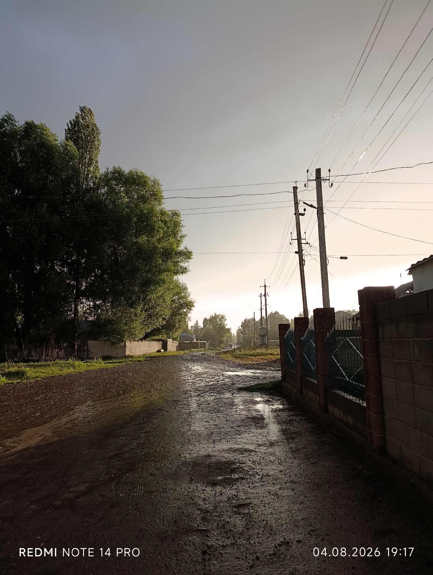 The image depicts a street in Kum-Döbä, Kyrgyzstan, after a rain shower. The dirt road is wet and reflects the sky, with puddles visible. On the left, a large, leafy tree dominates the foreground, with a low concrete wall and fencing behind it. Further down the street, smaller trees and buildings are visible. On the right, a brick wall with a green metal gate runs alongside the road, with utility poles and wires extending into the distance. The sky is overcast, with the sun partially visible on the horizon, casting a soft light. The overall impression is one of a rural or semi-urban setting experiencing inclement weather. A date and time stamp, "04.08.2026 19:17", and "REDMI NOTE 14 PRO" are overlaid on the image. There are no people or animals visible in the scene.