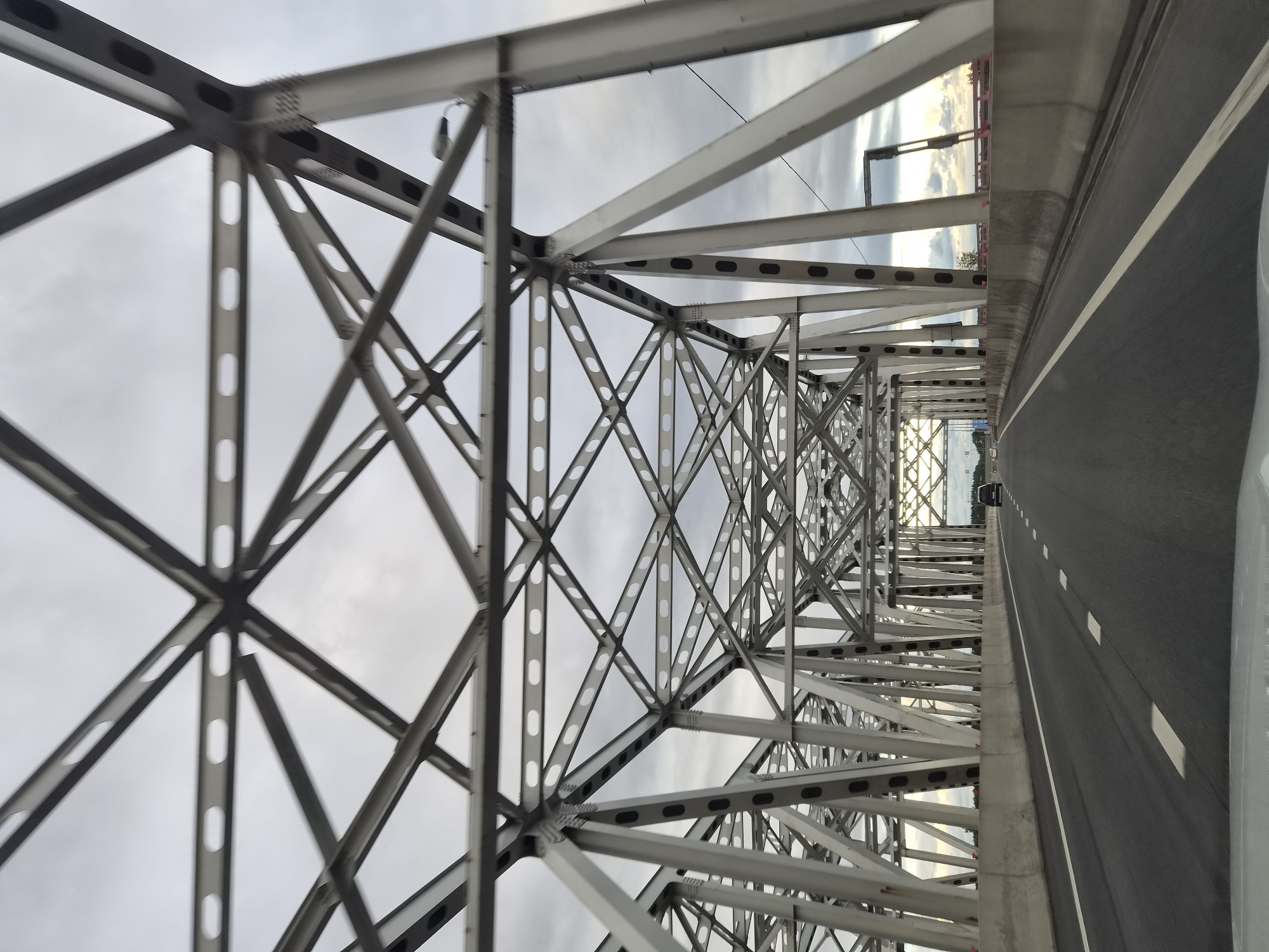 The image shows a view from a vehicle driving across a bridge. The bridge is constructed with a steel truss system, characterized by its intricate network of beams and supports. The roadway is asphalt, with clear lane markings. The sky above is overcast. The location is identified as Issad, Russia.