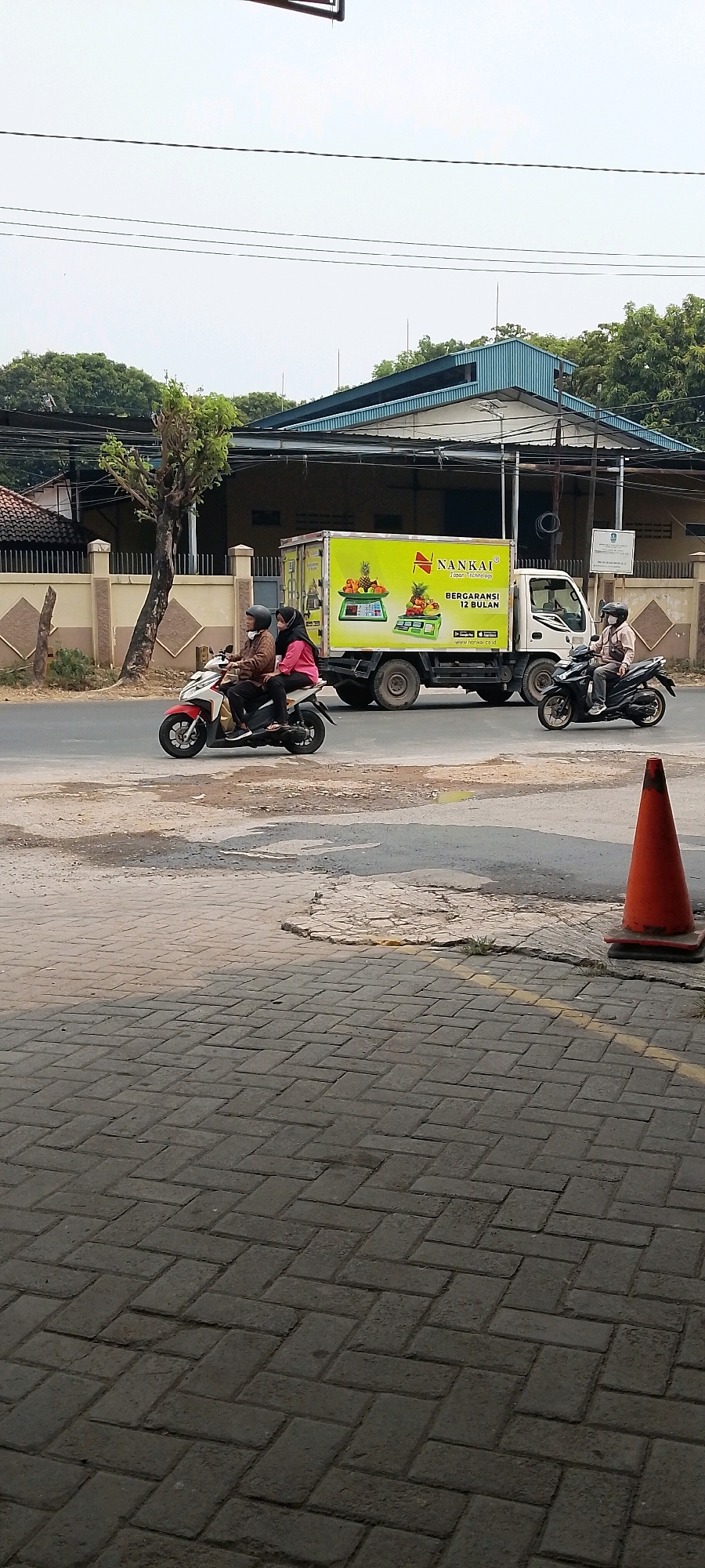 This image captures a street scene in Bekasi, Indonesia. A white truck with a bright yellow cargo box, featuring the "NANKAI" logo and an advertisement for fruits and vegetables, is parked on the right side of the road. The truck's side displays text in Indonesian, including "JAPAN TECHNOLOGY" and "BERGARANSI 12 BULAN" (12 Month Warranty). Two individuals are riding a red and white motorcycle in the left lane, heading away from the viewer. The rider wears a brown jacket and a black helmet, while the passenger wears a pink shirt and a headscarf, also with a dark helmet. Another motorcycle, ridden by a person wearing a light-colored jacket and a mask, is in the right lane, moving towards the right of the frame. An orange traffic cone is visible on the sidewalk in the foreground, adjacent to a patch of cracked asphalt and a paved walkway. In the background, a building with a blue, corrugated metal roof and beige walls is visible, with trees and power lines contributing to the urban landscape. The sky is overcast.