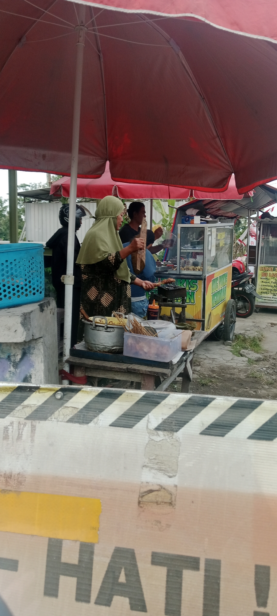 In Maguwoharjo, Indonesia, an outdoor food stall operates under a large red umbrella. A woman in a green hijab is preparing food, holding skewers of meat over a grill. She is positioned in front of a mobile food cart, which is decorated with yellow and green signage. Behind her, other individuals are visible, including one wearing a black helmet and another in the background. The food cart displays various items, including what appears to be cooked food in a glass-fronted display. In the foreground, a white barrier with black and yellow stripes is present, with the word "HATI!" (meaning "Careful!" in Indonesian) stenciled on it in large, dark letters. The ground is paved and appears somewhat weathered. A blue plastic basket is visible to the left of the scene.