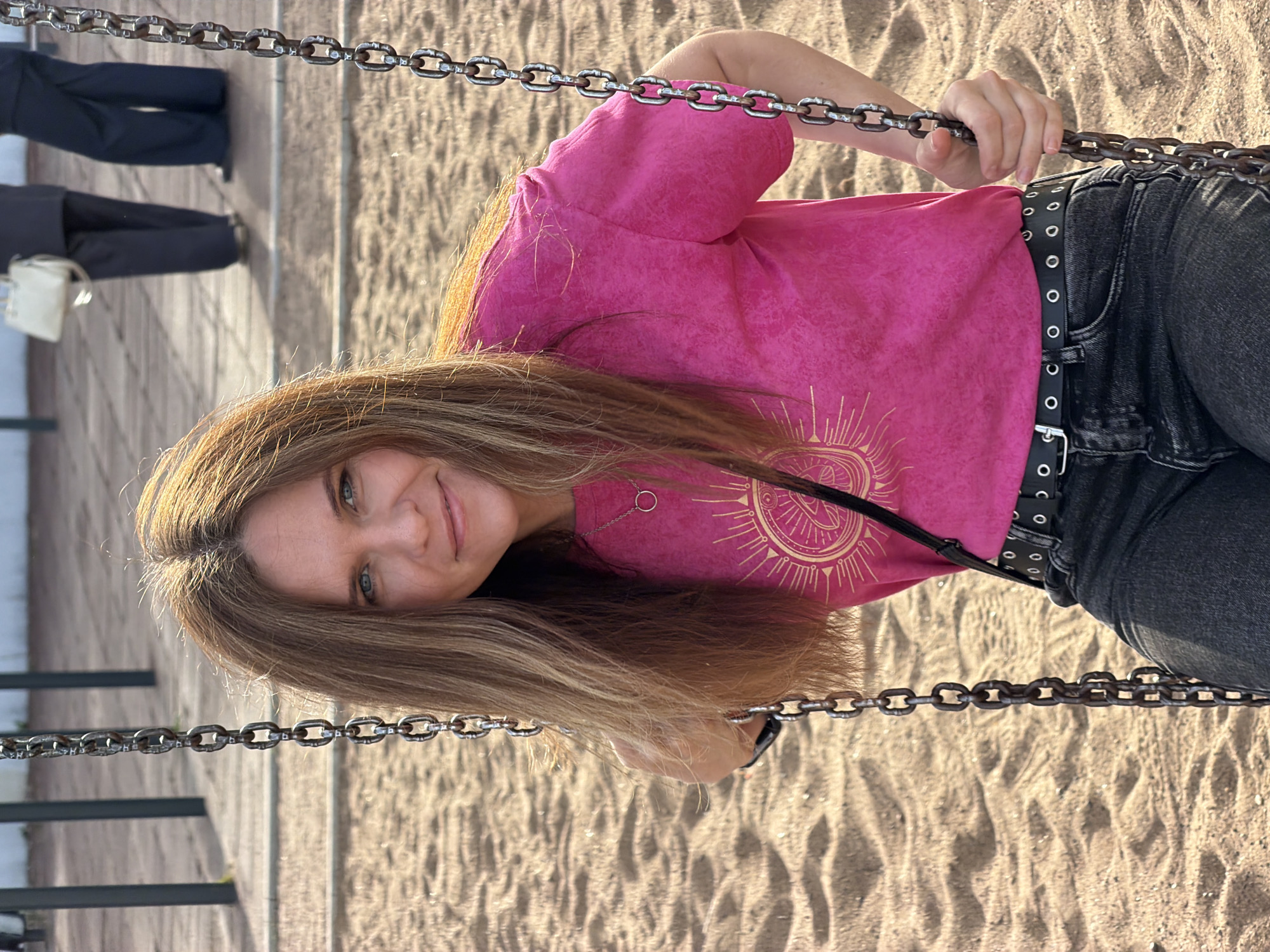 The image shows a woman on a swing in a sandy playground. She has long, blonde hair and is wearing a bright pink t-shirt with a gold sun design and dark jeans with a black belt. The woman is smiling and looking at the camera. The swing set chains are visible in the foreground and background. The sand on the playground is light brown and appears to be in soft, rippled formations. In the background, the legs of another person in dark pants and shoes can be seen, as well as a white bag hanging from their hand. The lighting suggests it is daytime, possibly late afternoon, with warm sunlight casting shadows. The overall mood appears cheerful and relaxed. No visible text is present in the image.