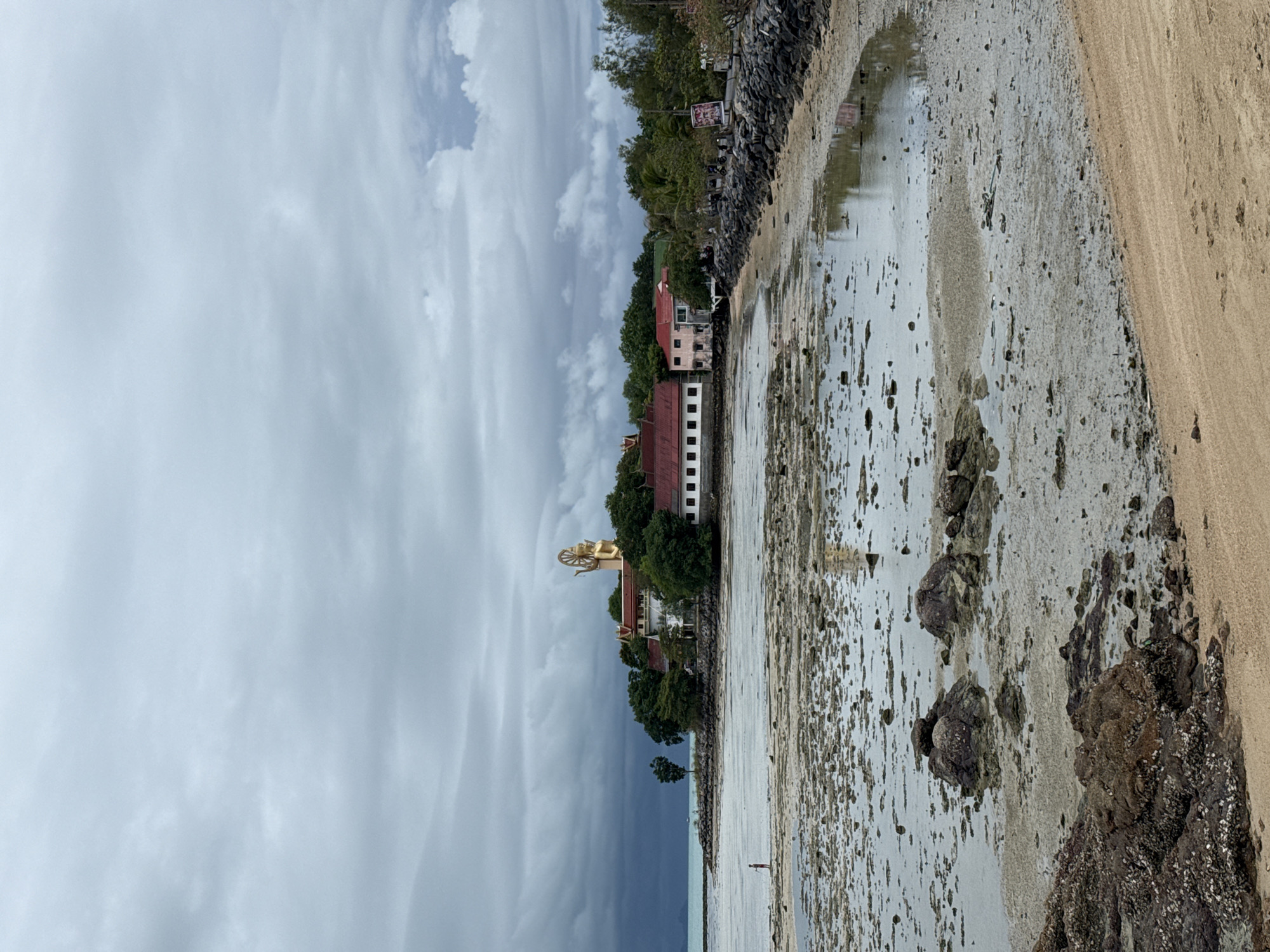 The image depicts a coastal scene in the Ko Samui District of Thailand, under a cloudy sky. In the distance, a large golden Buddha statue stands atop a hill, surrounded by lush green trees and buildings with red tiled roofs. The foreground is a rocky, sandy beach with tidal pools reflecting the overcast sky. A single person, appearing to be a child, stands near the water's edge in the middle ground. The overall atmosphere is one of subdued natural beauty, with a hint of human presence.