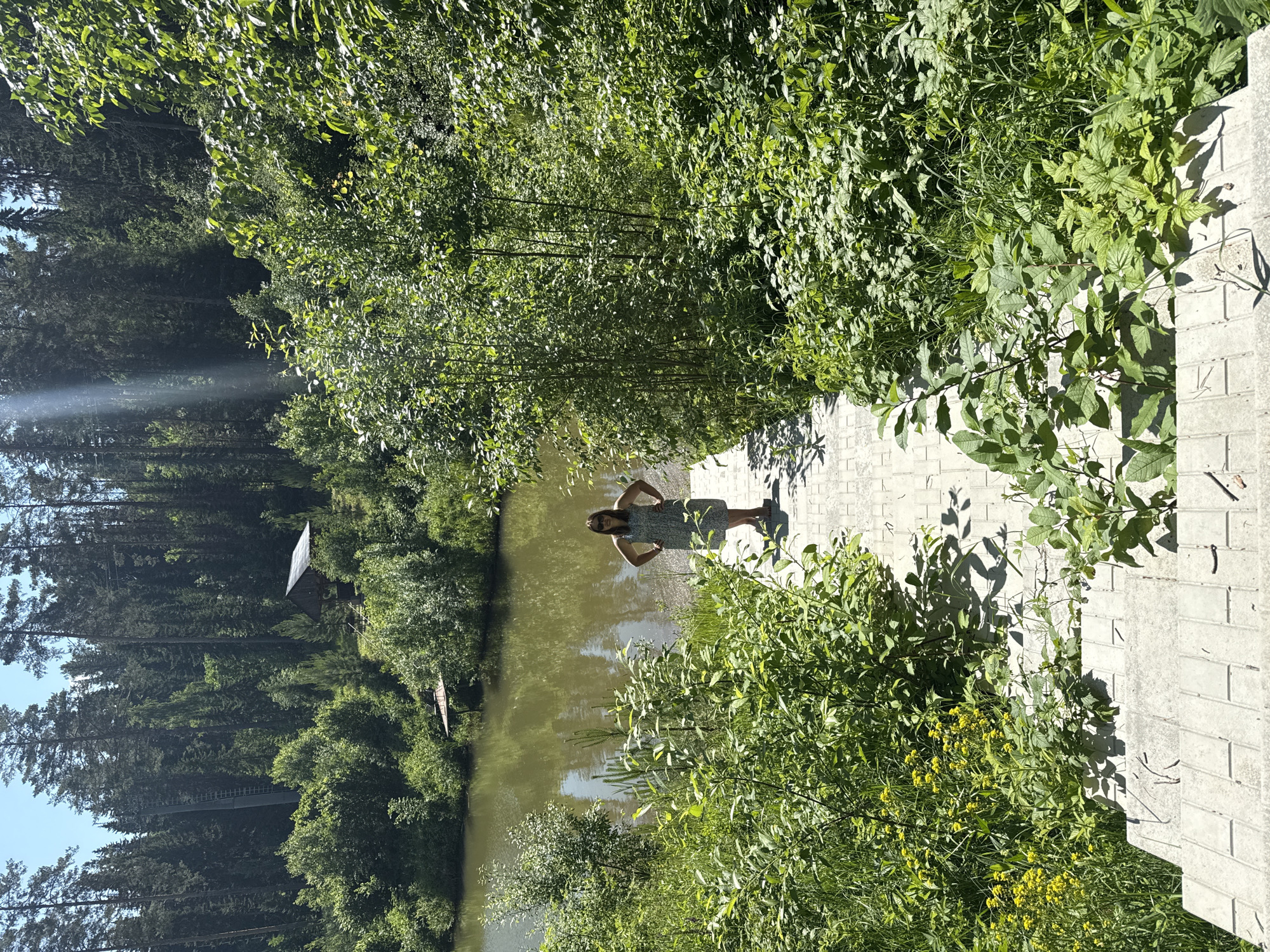 A woman stands on a tiled walkway leading down towards a lake. She is wearing a patterned dress and sunglasses, with her hands on her hips. The walkway is surrounded by lush green vegetation. In the background, a dense forest of tall trees borders the lake. The sky is clear and blue with a few sunbeams filtering through the trees. There is a small wooden structure visible in the distance amongst the trees on the opposite bank of the lake. The scene suggests a natural, serene outdoor setting, possibly a park or a recreational area near Makarovo, Russia.