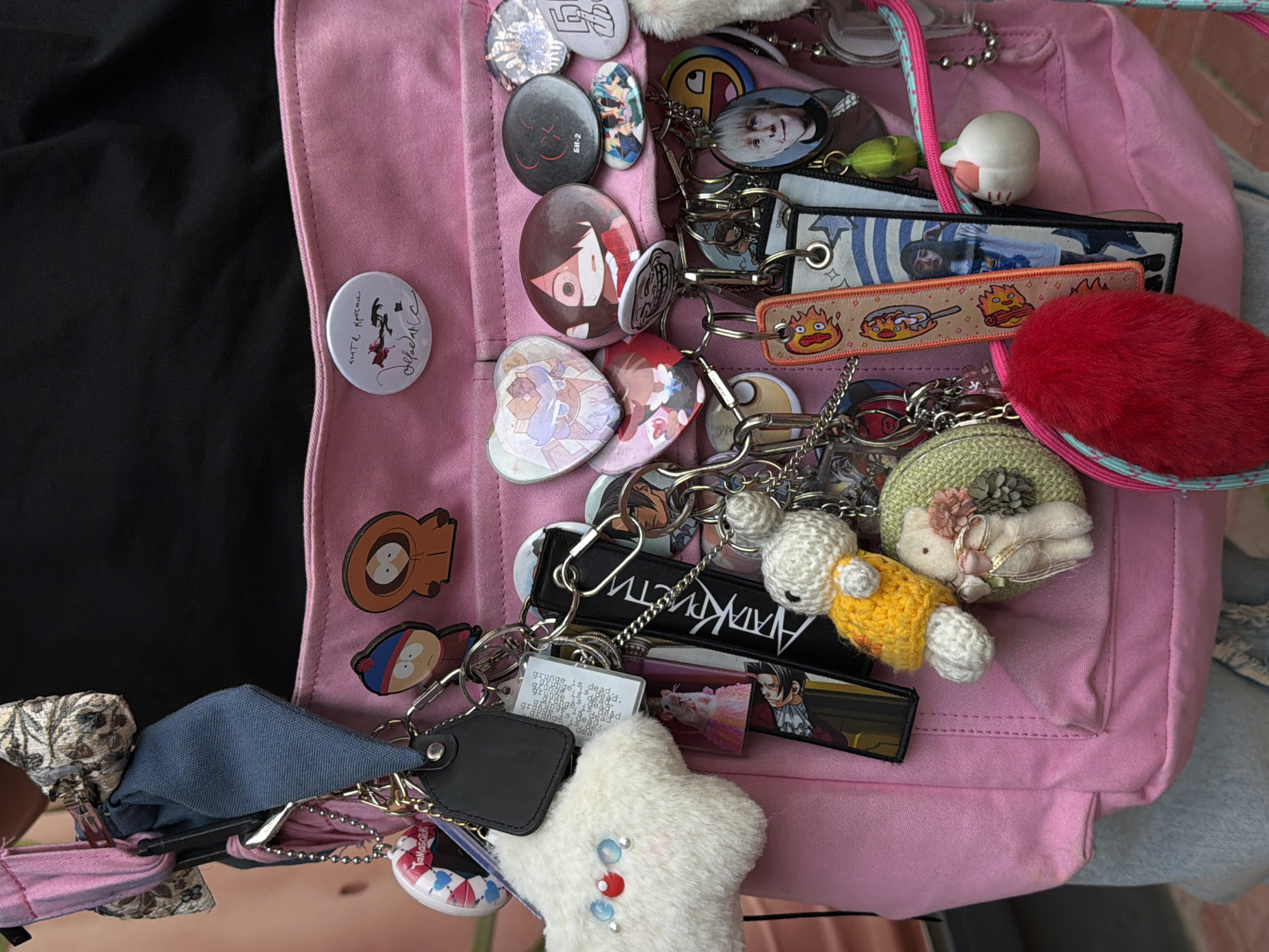 The image shows a light pink backpack adorned with various pins, keychains, and small plush ornaments. The pins include images of anime characters, a rock band named “БИ-2”, and cartoon characters from the show “South Park.” The keychains are diverse, with themes from video games like “Ace Attorney”, anime like “Howl’s Moving Castle”, and general decorations like a white fuzzy star. A few plush figures are attached, including one knitted with yellow yarn, and a red fuzzy ball. The backpack is partially obscuring a black garment on the left and some lighter-colored clothing on the right. The background is indistinct, making it impossible to determine if the image was taken in Budva, Montenegro, or any other specific location.