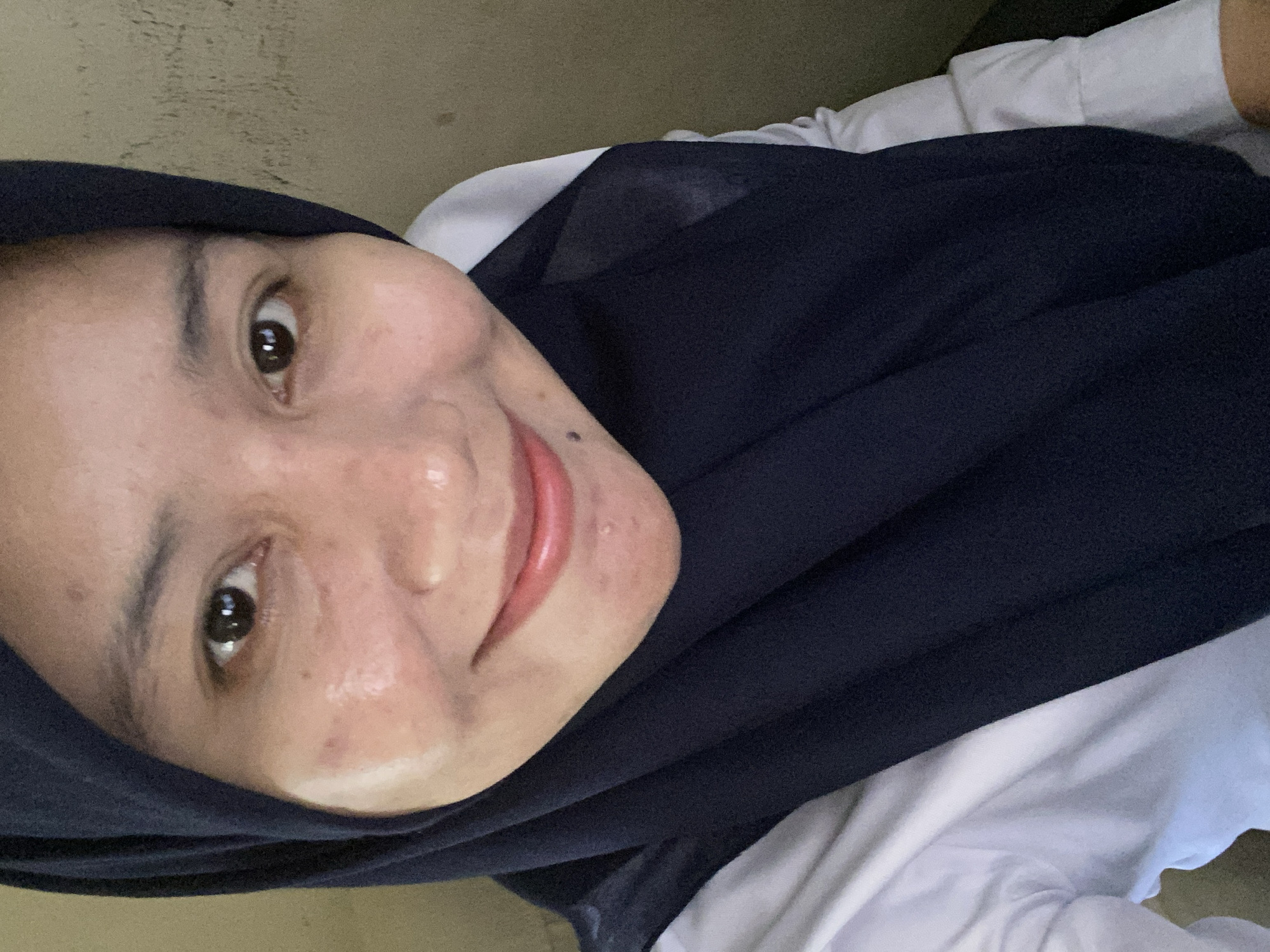 A medium shot selfie captures a young woman wearing a dark blue hijab and a white collared shirt. She is looking directly at the camera with a gentle smile, her eyes are dark brown. Her skin appears slightly oily. The background is a textured, beige wall. The image quality is moderate, with some blurriness and graininess. The scene is indoors, likely in a residential or office setting within Banjarbaru, Indonesia. There are no other people or objects of significance in the frame. The lighting is soft and diffused, suggesting natural light from a window or an overhead light source.