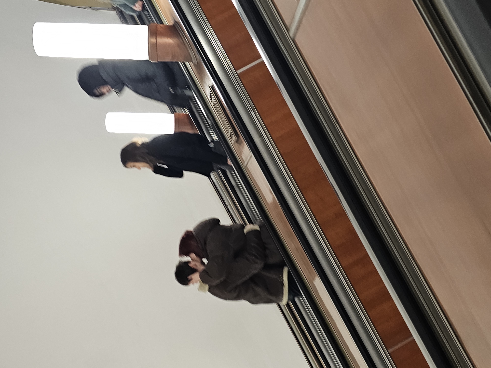 People ride an escalator inside a metro station. The location is likely in Moscow, Russia, given the interior architectural style of the station. Three individuals are visible in the frame. The closest person is wearing a dark jacket. The second person is wearing a black coat. The third person is wearing a black jacket. Bright lights illuminate the escalator.