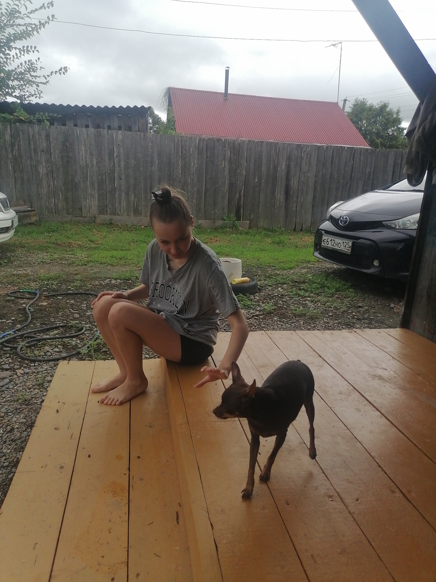 In Novopokrovka, Russia, on a cloudy day, a young female with her hair in a bun sits on a wooden porch. She wears a grey t-shirt with "BROOKLYN" printed on it and black shorts. Her bare feet are visible as she rests her knees. A small black dog stands on the porch in front of her, facing left. The porch is made of light-colored wooden planks. Behind the porch, a wooden fence runs horizontally, with a red-roofed building and trees visible beyond it. A black car is parked in the background to the right of the fence. A garden hose is visible on the ground to the left of the porch.
