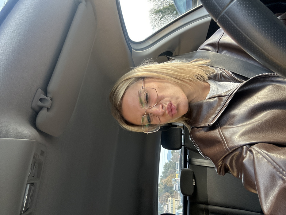 A blonde-haired female, wearing a dark brown zippered jacket over a light-colored top and large square-framed glasses, is seated in the driver's seat of a vehicle. She is taking a selfie, making a "duck face" expression with pursed lips and a slightly squinted right eye. A seatbelt diagonally crosses her chest. The car's interior features a light-colored headliner with an overhead light fixture and a sun visor, and dark-colored rear seats are visible behind her. A black steering wheel is partially visible on the right side of the frame. Through the rear window, a commercial building with a sign displaying "WORK" and partially visible text (likely "AT WORK AUTO") is visible. This business is situated at 505 Boulevard des Ferrayolles in Villeneuve-Loubet, France. A road or parking area in front of the building shows other vehicles, including a white truck. In the distant background, tree-covered hills or mountains are present under a bright sky. To the right of the vehicle, bare tree branches and bright sky are visible through a side window.