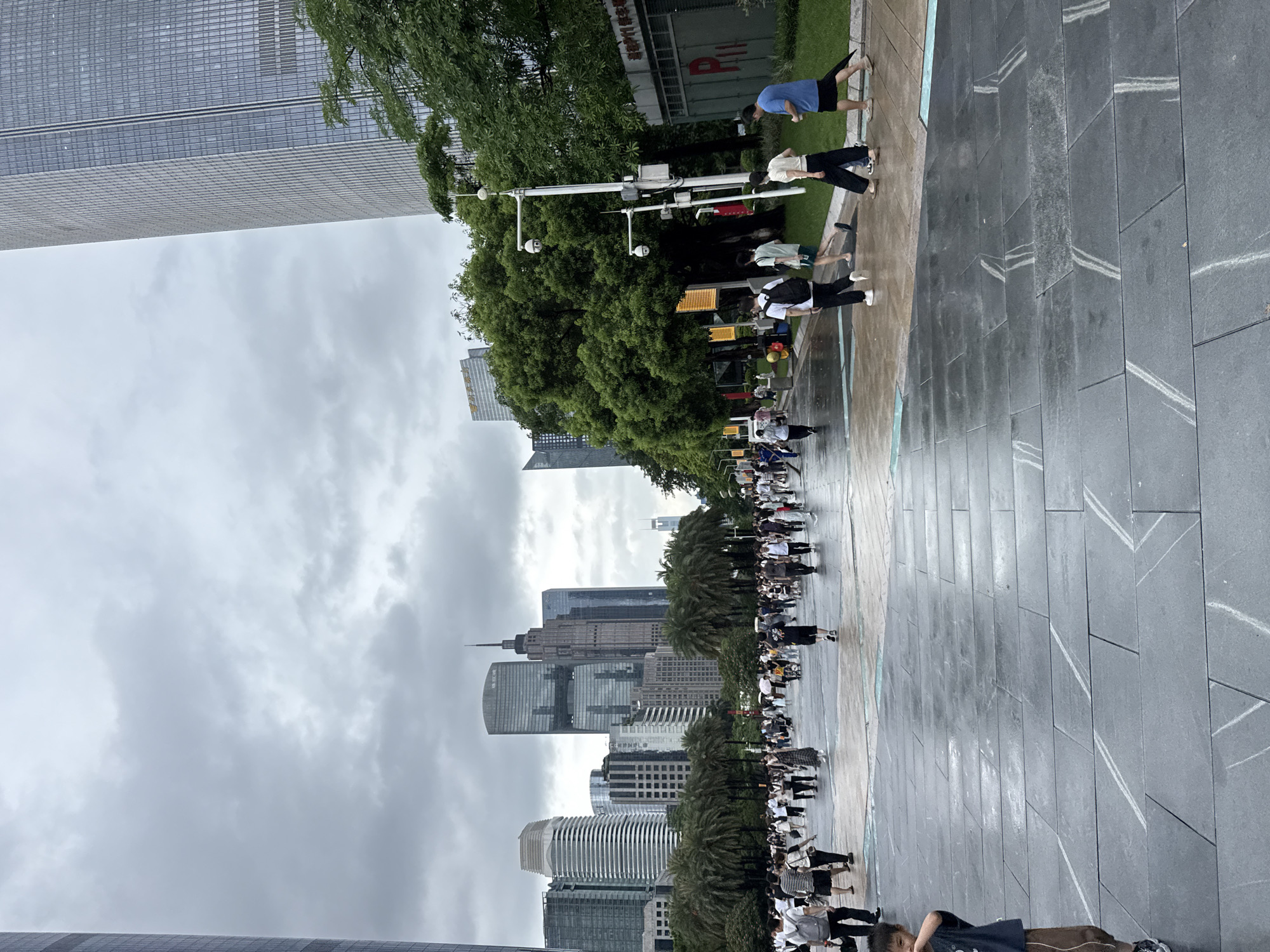 The image depicts a public square in Tianhe District, China, under a cloudy sky. A wide, wet paved area, possibly from recent rain, dominates the foreground. Several large, modern skyscrapers with glass facades form the backdrop of the scene. A crowd of people is walking across the square, some individually, others in small groups. There are trees and palm trees lining the sides of the square. Several CCTV cameras are visible on a pole to the right of the image.