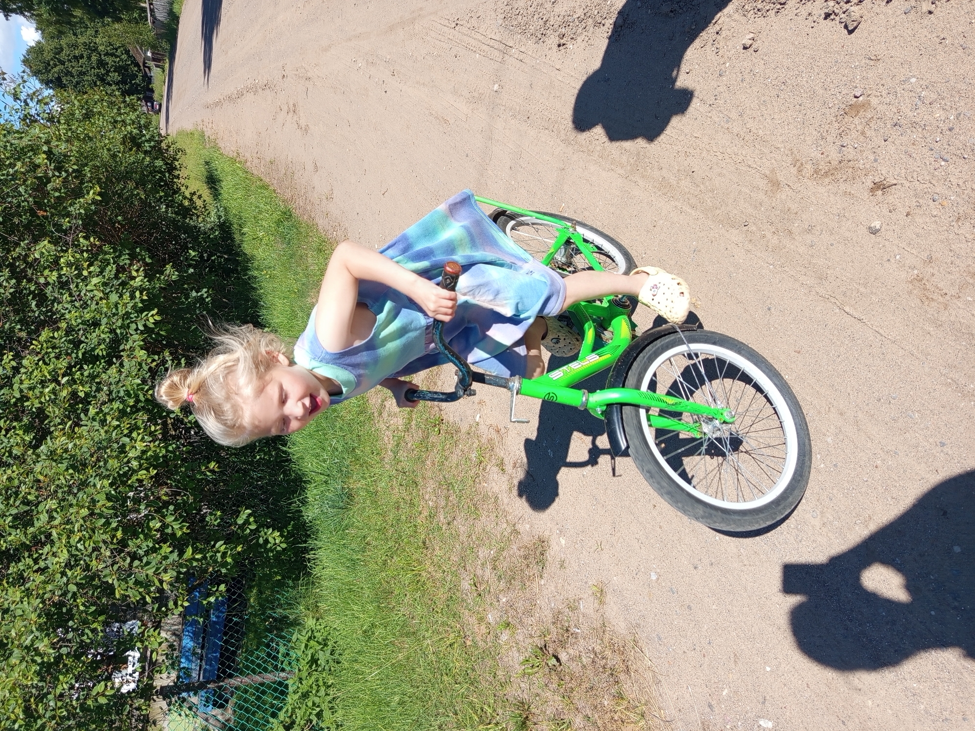 In Ostrovtsy, Russia, a young blonde girl with her hair in a bun is pictured on a bright green bicycle. She is wearing a tie-dye dress in shades of blue and green, and light-colored clogs. The girl is leaning forward, with her tongue sticking out slightly, appearing engaged with the bicycle. She is holding onto the handlebars with one hand.
The bicycle has a visible chain and spoke wheels with white rims. The scene is outdoors on a sandy or dirt path, with green grass and bushes bordering the path. In the background, there are trees and hints of structures, suggesting a rural or semi-rural environment. The sun is bright, casting shadows on the ground, indicating it is daytime, likely on a clear day. The overall mood seems playful and active, with the girl enjoying herself outdoors.