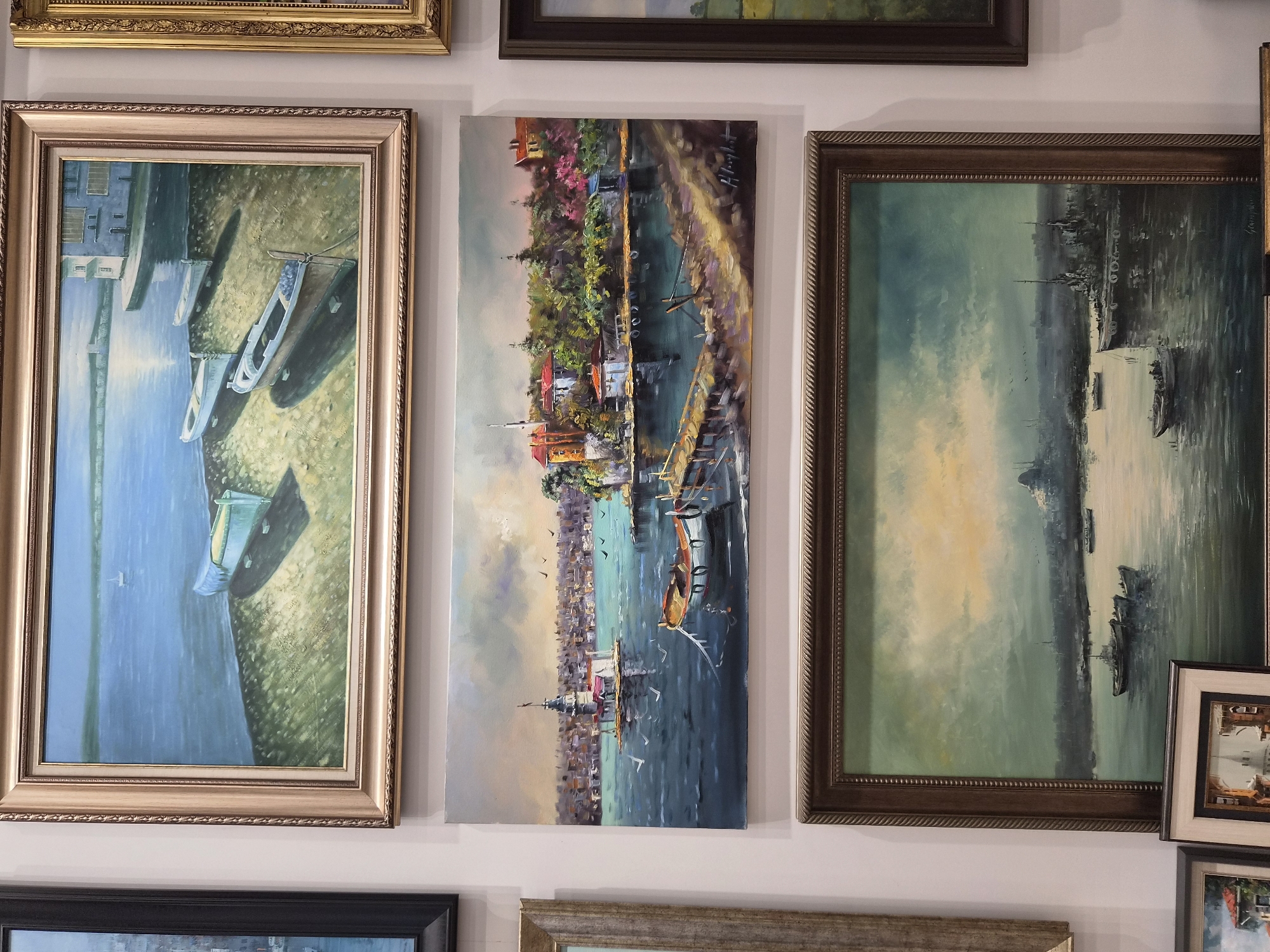 The image displays three framed oil paintings hanging on a white wall. The topmost painting depicts a tranquil seascape with boats beached on a sandy shore, a distant building, and a calm expanse of water. The middle painting is a vibrant cityscape with a prominent tower on an island, boats in the foreground, and a detailed waterfront with lush greenery and houses. This painting is signed by H. Kayamart. The bottom painting presents a broad view of a body of water with several boats, including a large ship, and a distant city skyline under a cloudy sky with hints of sunlight. The location of Maltepe, Turkey, is not explicitly depicted or indicated within the visual content of the image.