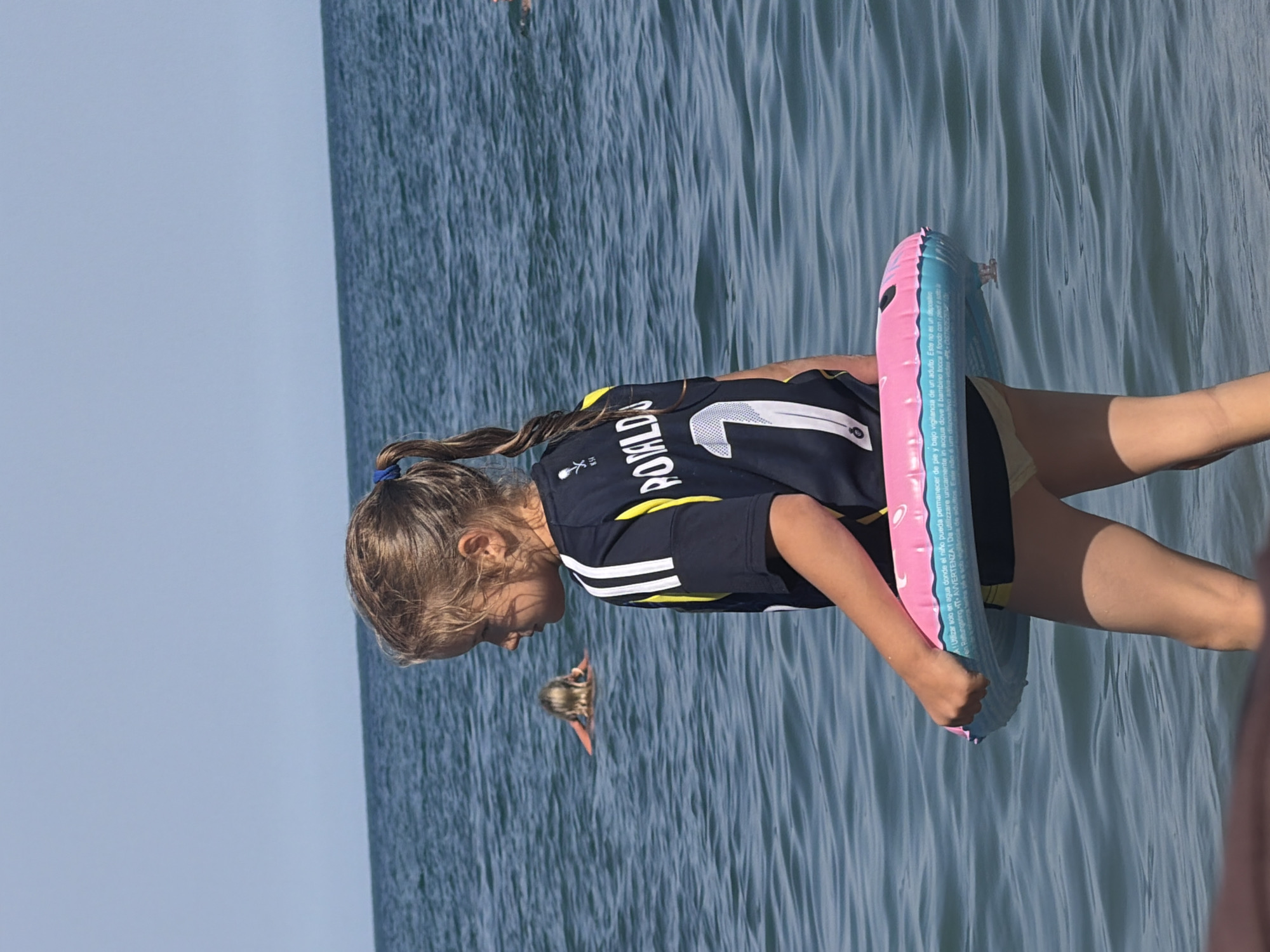 This media file shows a young girl standing in shallow water, likely at a beach or lake. She is wearing a dark blue t-shirt with yellow and white stripes on the sleeves and a white design on the back with the number "7" and the name "POMLD" visible. She has her hair in a braid and is holding a pink and blue inflatable ring. Another person, partially visible, is also in the water further away. The water is a deep blue-green with visible ripples, and the sky is a clear light blue, suggesting a sunny day, likely in the morning or afternoon. Text is visible on the inflatable ring, including "A! es un" and "AVVERTENZA! Da utilizzare unicamente in acqua dove il bambino tocca fondo conpleksot". The setting appears to be a body of water, possibly Partenit, Ukraine. The overall mood seems relaxed and summery.