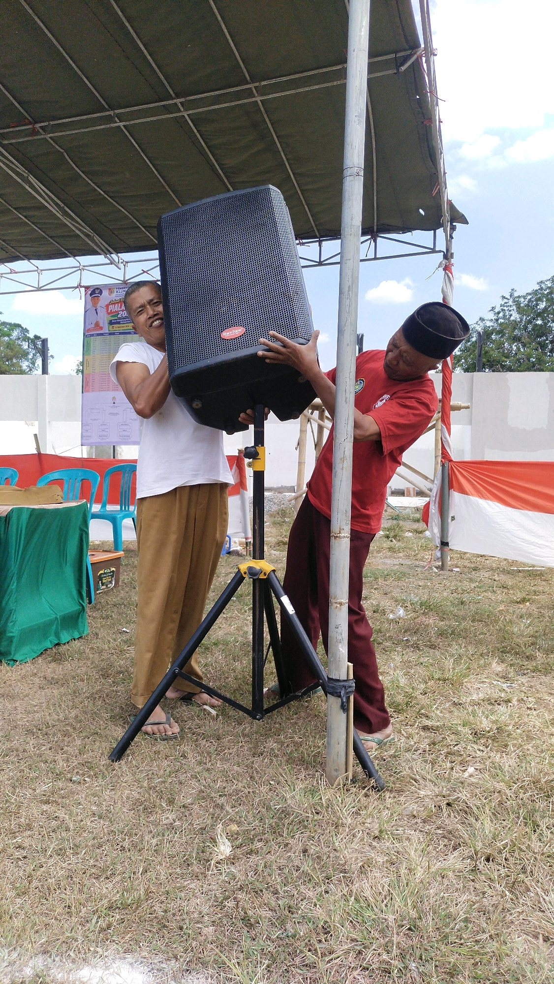 Two men are working outdoors to set up a large speaker on a tripod stand. The man on the left, wearing a white t-shirt and tan pants, is looking at the camera with a wide smile as he holds the speaker. The man on the right, wearing a red shirt and maroon pants with a black cap, is holding the speaker and adjusting its position. The speaker is black and has a brand name "Han-Tek" on it. The tripod stand is black with yellow accents. In the background, there is a green tablecloth draped over a table, blue plastic chairs, a white wall, and red and white Indonesian flags. The scene appears to be in a grassy outdoor area, possibly a public event space. The presence of Indonesian flags and the mention of Klungkung, Indonesia, suggests this location is likely in Klungkung, a regency in Bali, Indonesia. The overall atmosphere is one of preparation and cooperation for an event.