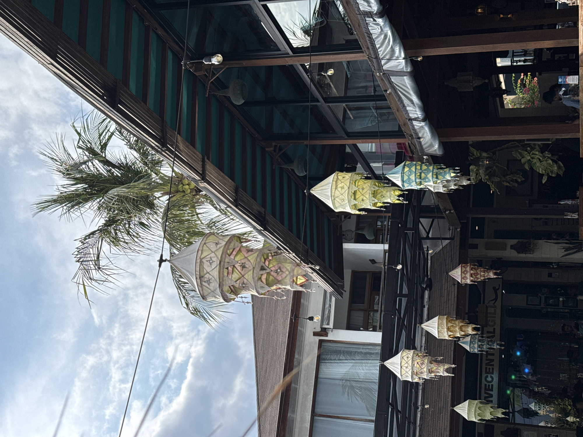 This outdoor shot, taken from a low angle, features hanging lanterns with decorative patterns. The lanterns, strung along a dark metal framework, are under a building's awning. A palm tree is visible on the left side of the frame, and a sign reading "IVECENTRAL GIL" is partially visible below one of the lanterns. The sky appears cloudy. The image was taken in Unknown, Indonesia.