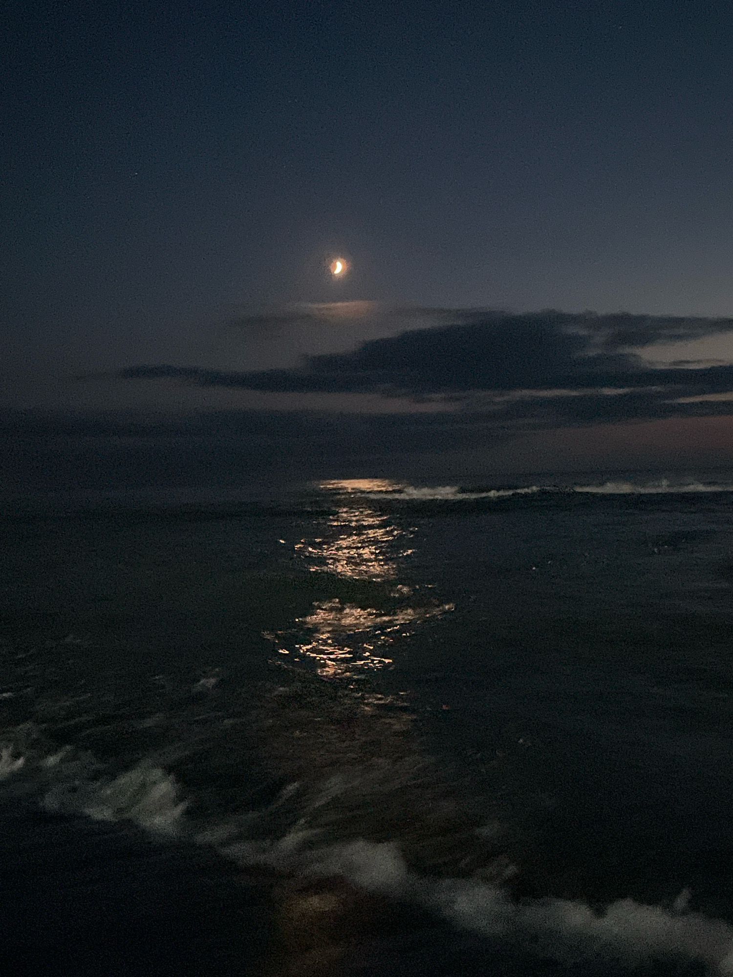 The image shows a dark seascape under a twilight sky with a crescent moon. The moon is visible in the upper central portion of the frame, casting a shimmering reflection on the ocean's surface. Dark clouds are present in the sky, obscuring some of the moonlight. Gentle waves are breaking on the shore in the foreground. The scene suggests the location is a beach or coastline at night. Ureki, Georgia is known for its Black Sea coastline and magnetic sand beaches, which are often visited at various times of day and night.