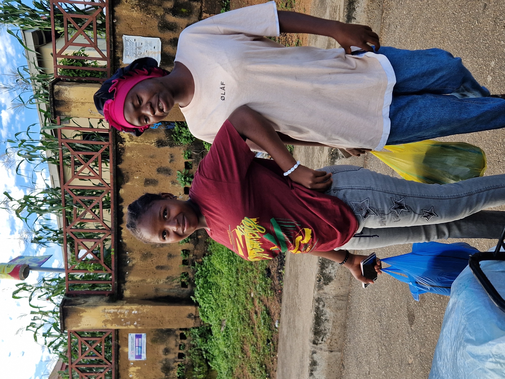 Two young Black females are standing outdoors on a paved surface next to a brown, stucco wall with a red decorative railing and greenery growing behind it. Corn stalks are visible above the wall against a cloudy blue sky. The female on the left has dark skin and braided hair pulled back. She is wearing a maroon t-shirt with a graphic design and light-colored jeans with star embellishments. She is holding a smartphone in her left hand and a blue plastic bag in her right. The female on the right has dark skin and is wearing a light-colored t-shirt with "ØLÅF" printed on it, and blue jeans. She has a pink headwrap and black bonnet covering her hair. She is holding a yellow plastic bag in her right hand. The location is identified as Kabong, Nigeria, in the city of Jos.