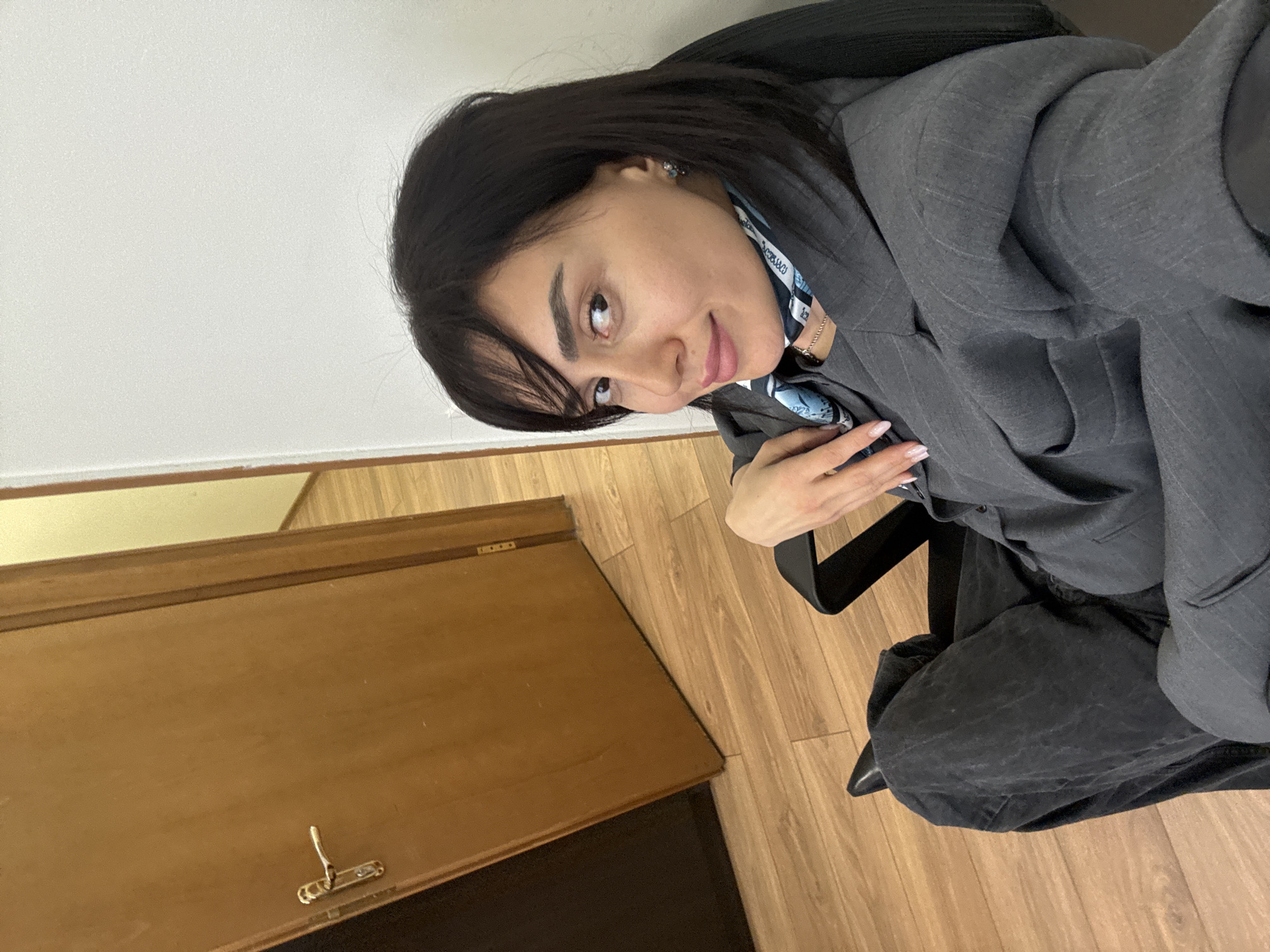 A woman with dark hair is seated in an office chair, wearing a gray blazer and a blue and white patterned scarf. She is smiling and looking toward the camera. The background includes a white wall and a wooden door set within a room with wooden flooring. The location is likely indoors in Tbilisi, Georgia.