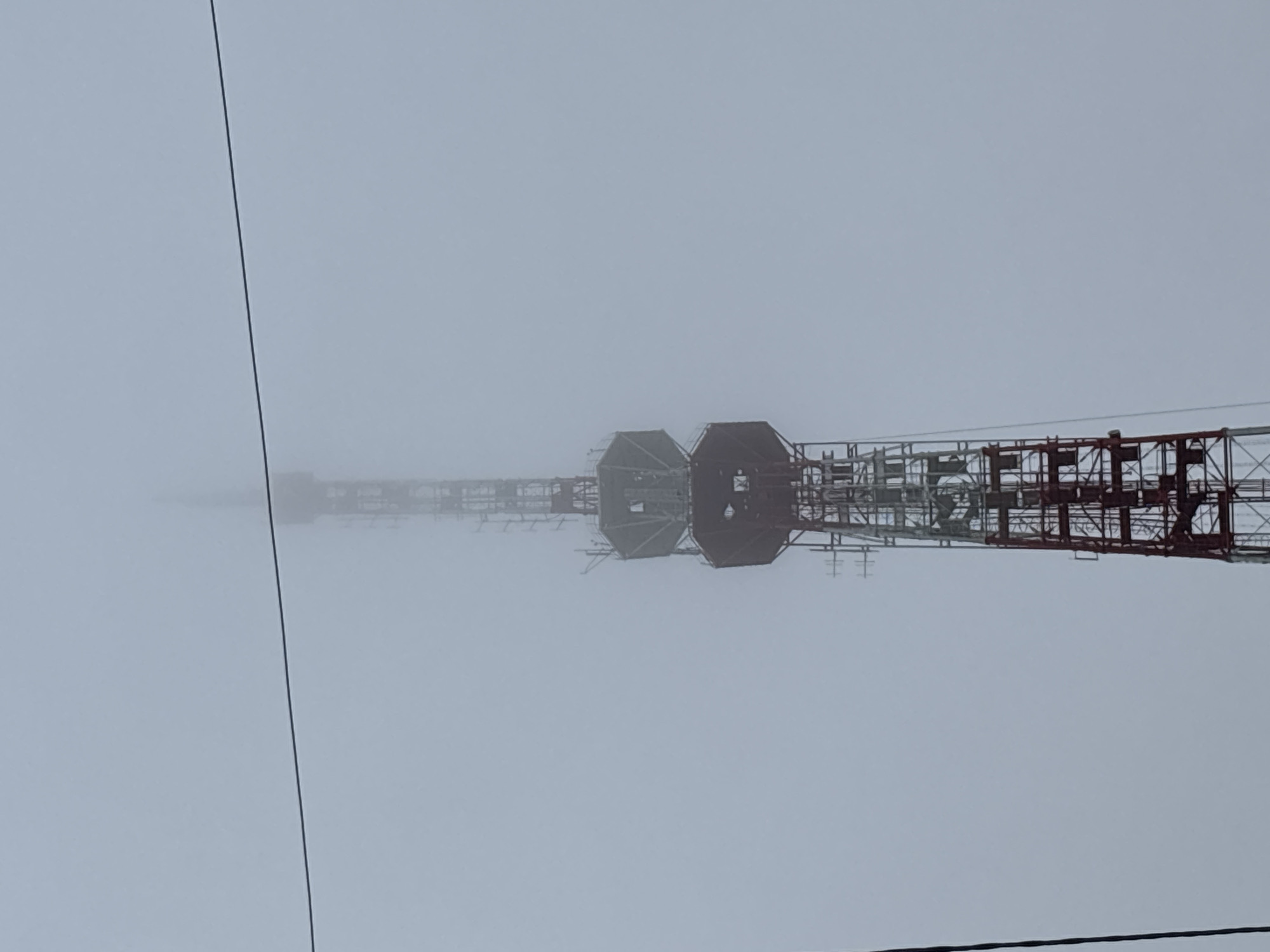 The image shows a close-up view of a tall, red and white lattice tower, likely a transmission tower, reaching into a foggy sky. The tower has several levels with platforms and antennae, and its top portion is obscured by the fog. A thin black wire stretches horizontally across the upper portion of the frame. The scene is dominated by the muted tones of the fog, creating a somber and slightly mysterious atmosphere. The tower appears to be in an industrial or open area, given its scale and the lack of surrounding buildings visible. While the exact location within Blagoveshchensk, Russia, cannot be precisely identified from the image alone, such towers are often situated on the outskirts of cities or in designated communication zones. There are no people or other discernible objects in the immediate vicinity of the tower, emphasizing its isolation within the foggy environment.