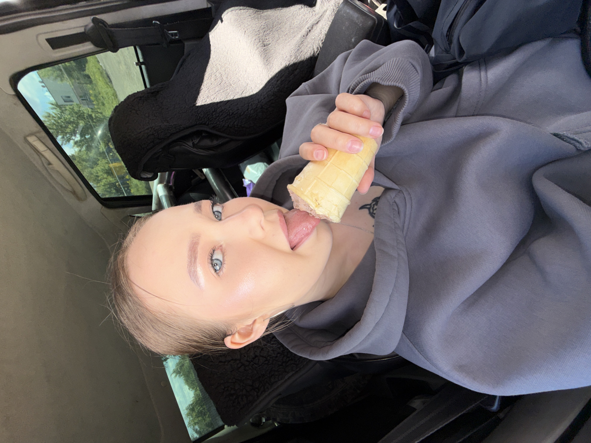 The image shows a woman with light skin and blonde hair in a car, eating an ice cream cone. She is wearing a gray hoodie. Through the car window, trees and a building can be seen. The location of the scene is in the city of Фоки, Russia.