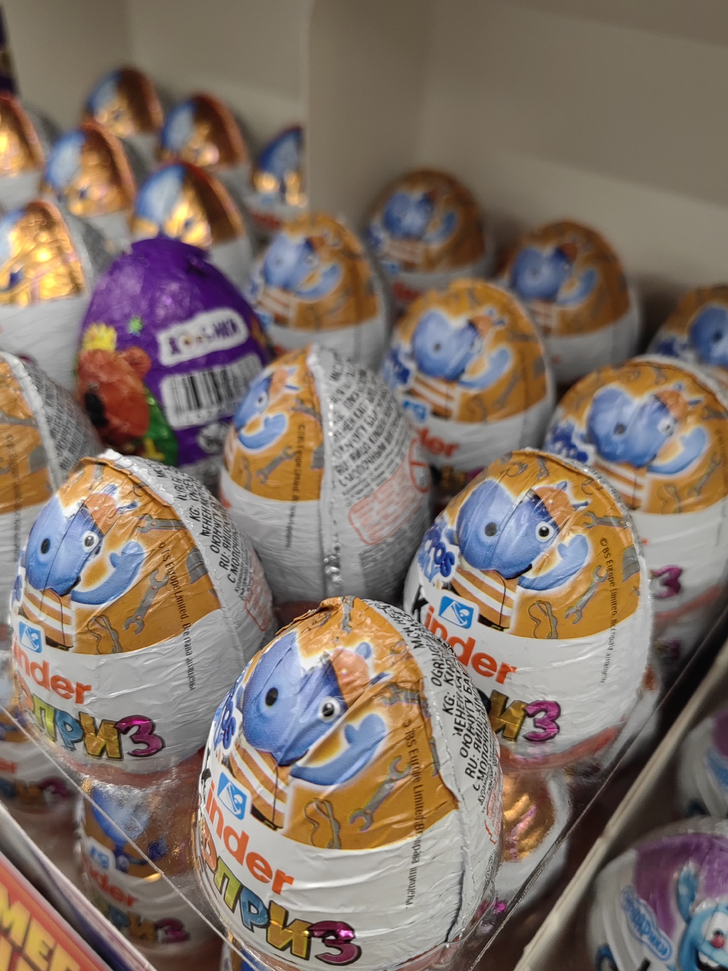 A close-up, eye-level shot shows a display of Kinder Surprise eggs in a retail setting. The eggs are individually wrapped in foil, with most featuring a blue cartoon character on an orange and white background. Some eggs are partially obscured by others. In the foreground, one egg has the word "Kinder" printed in red, with "ПРИЗ" in iridescent pink and yellow letters below it. Behind this, another egg has similar branding. To the left, a purple-wrapped egg with a red character is visible. The background consists of more rows of these chocolate eggs. The lighting is bright, suggesting an indoor environment such as a supermarket or convenience store. The image appears to be taken in a city in an unknown location.