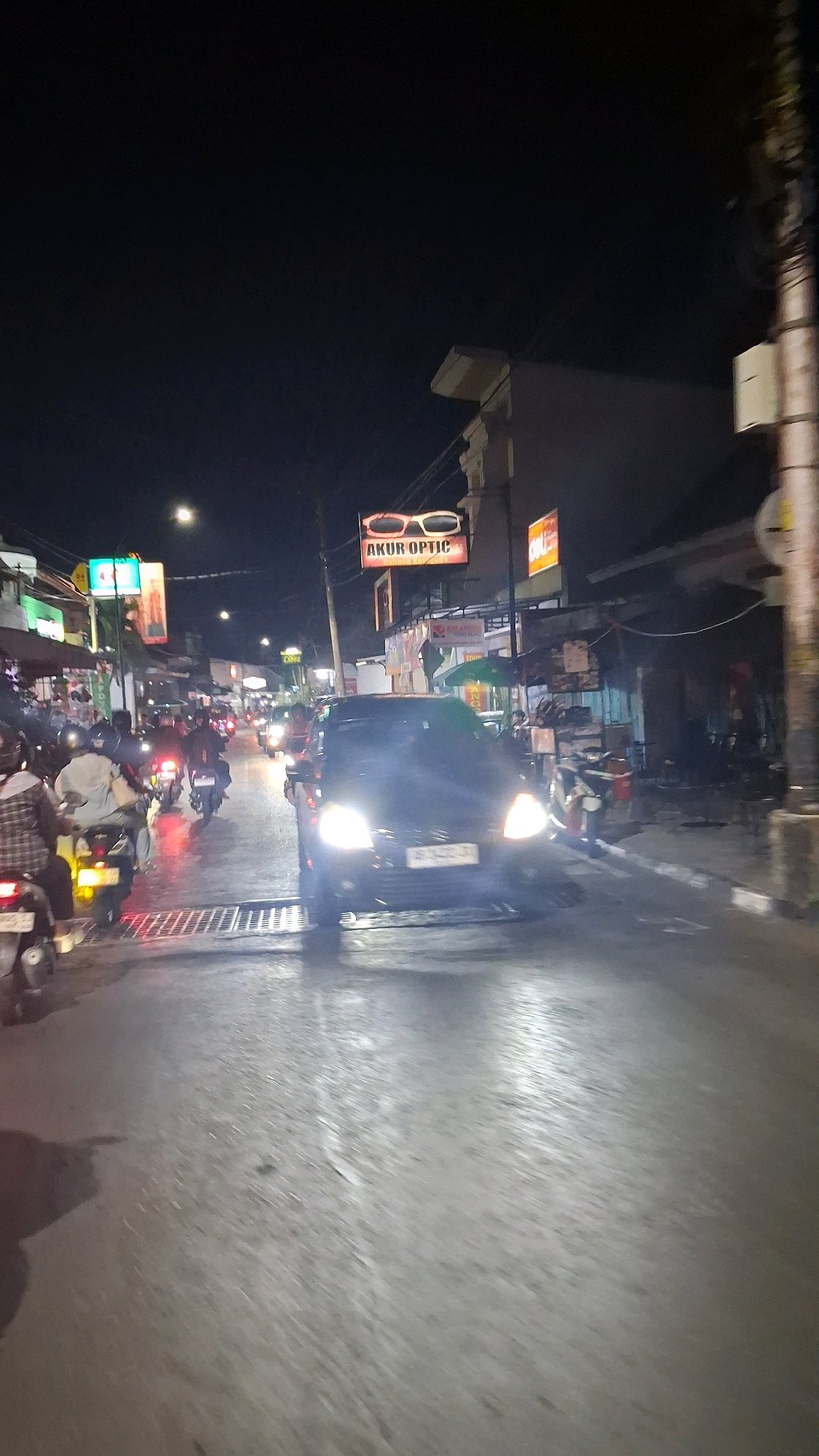 A vertical, night-time, motion-blurred photograph depicts a street scene in Yogyakarta, Indonesia, likely during a period of moderate traffic. In the foreground, the wet asphalt road surface reflects the glow of headlights and streetlights, creating a streaky, luminous effect. A dark-colored car, its headlights illuminated, occupies the center of the frame and is moving away from the viewer. To the left, several motorcycles with their riders are also visible, heading in the same direction.
Along the sides of the street, shopfronts and stalls are illuminated with neon signs and bright lights. A prominent sign, featuring a pair of stylized glasses, reads "AKUR OPTIC". Other shop signs are also visible but are less distinct due to the motion blur and low light conditions. The buildings appear to be of varying heights and architectural styles, typical of a commercial street in an Indonesian city. The overall impression is that of a busy urban environment at night, with constant movement of vehicles and activity from the surrounding businesses. The presence of water on the road suggests recent rain or a damp evening.