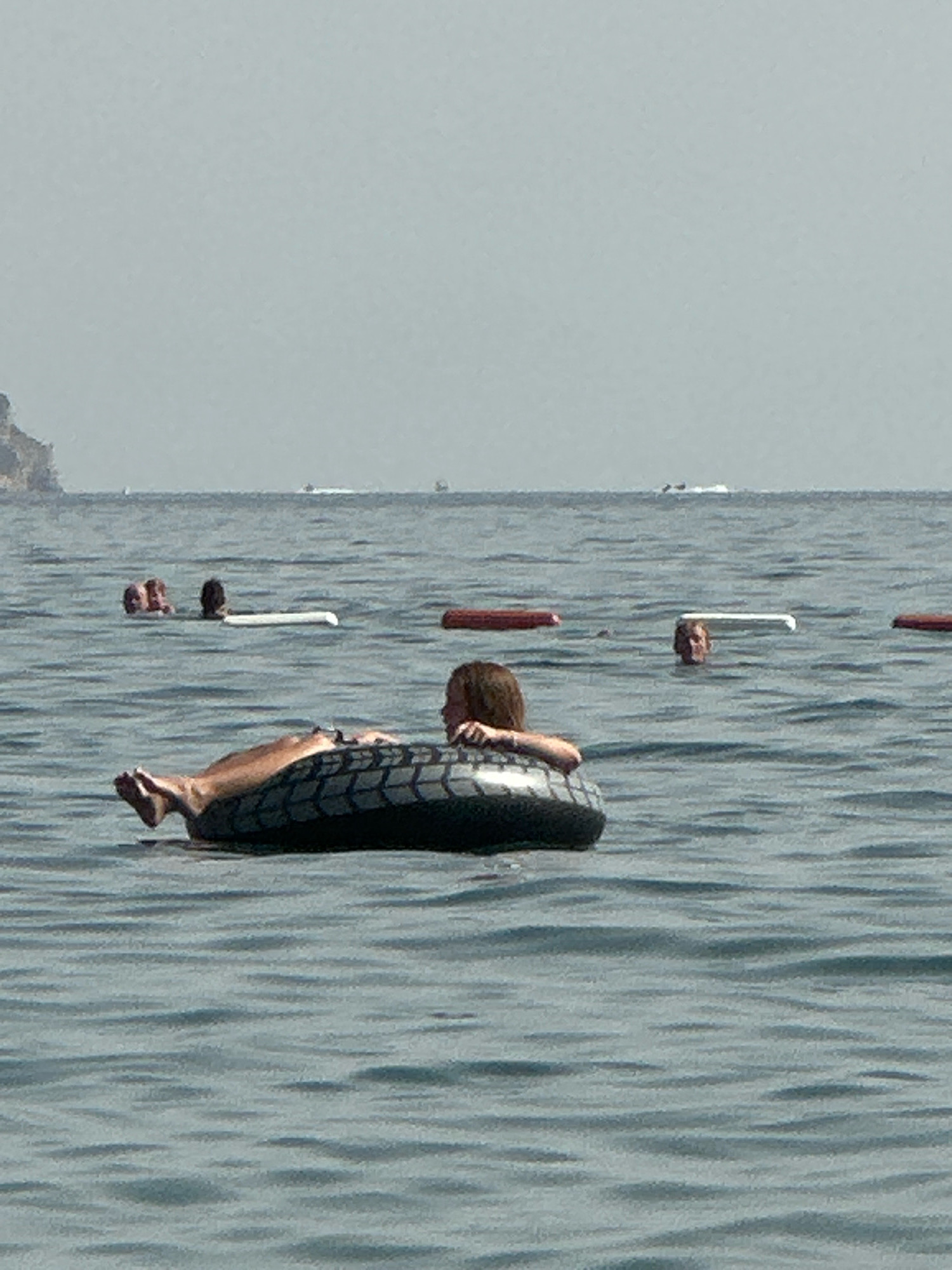 A person is floating on their back in the sea, supported by a tire inner tube. Their legs are extended out of the water, and their arms are resting on the inner tube. The person's head is tilted back, and their hair is visible. The water is calm and blue, with some gentle ripples. In the background, other people are visible in the water, some with their heads above the surface. There are also some buoys or markers in the distance. The sky is hazy and light blue. The location is the sea in Boreti, Montenegro.