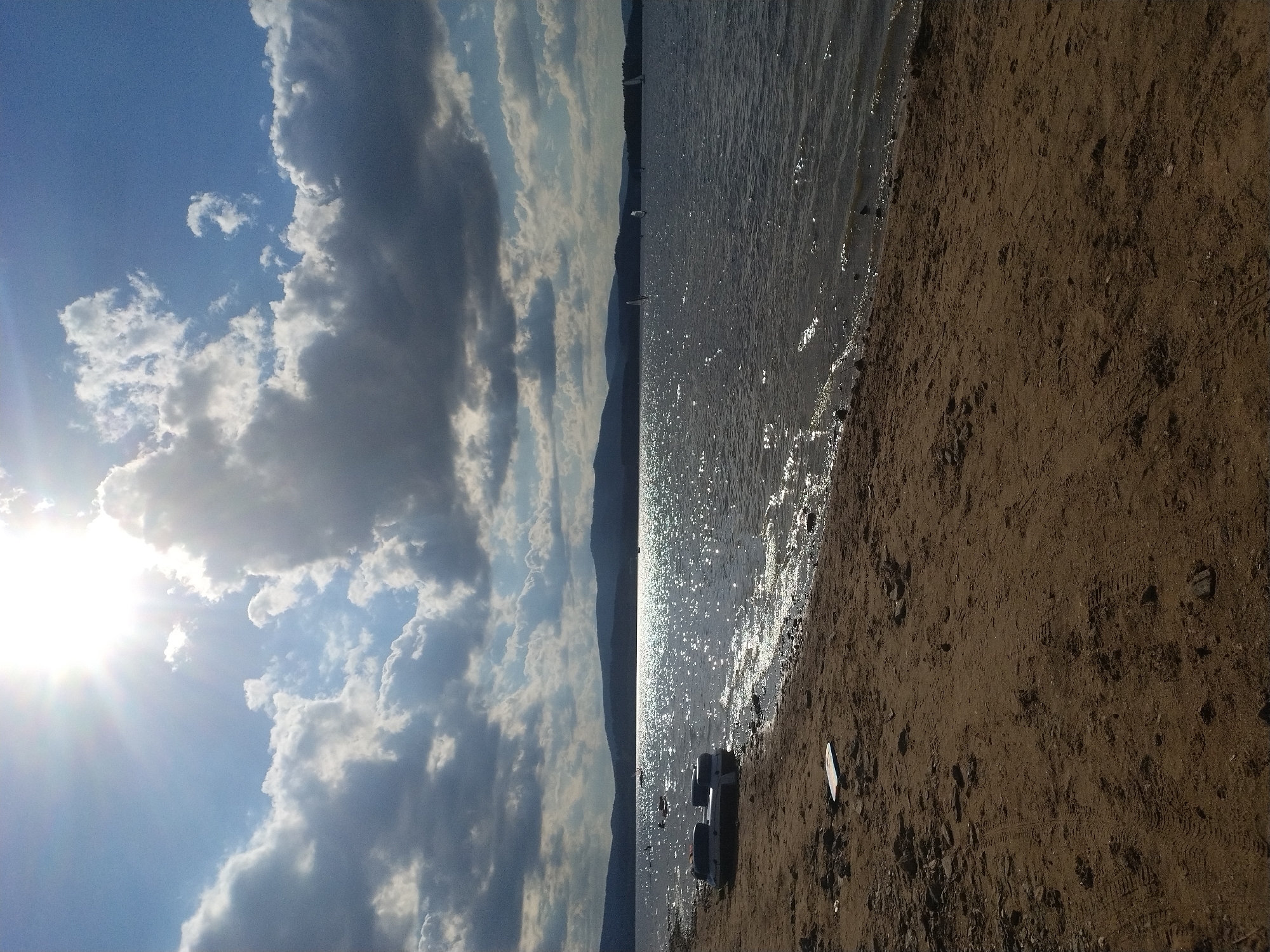 The image depicts a sunlit lake scene with a sandy shore. The sun is bright and high in the sky, casting a glare on the water and creating lens flare. Clouds are present in the sky, some are dark and appear to be rain clouds, while others are bright white and illuminated by the sun. The lake is calm with ripples on the surface, reflecting the sunlight. On the shore, there is an inflatable boat or raft pulled up onto the sand. In the distance, a line of hills or mountains can be seen under the clouds. Several sailboats are visible on the lake. The sandy shore in the foreground has tire tracks and scattered rocks and debris. The image does not show any people. The location is the Radslav Reservoir, near the village of Radslav in the Czech Republic.