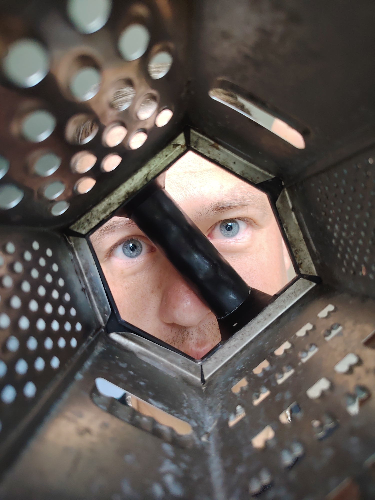 The image, taken in Chaikovsky, Russia, shows a close-up perspective through a grater. A man's face is visible, with his blue eyes looking directly at the camera. His right eye is the most prominent feature, with the iris clearly discernible. A portion of his nose and upper lip are also visible. He appears to be holding the grater. The grater itself is made of metal and has numerous holes of various shapes and sizes. The background is out of focus, but appears to be an indoor setting.