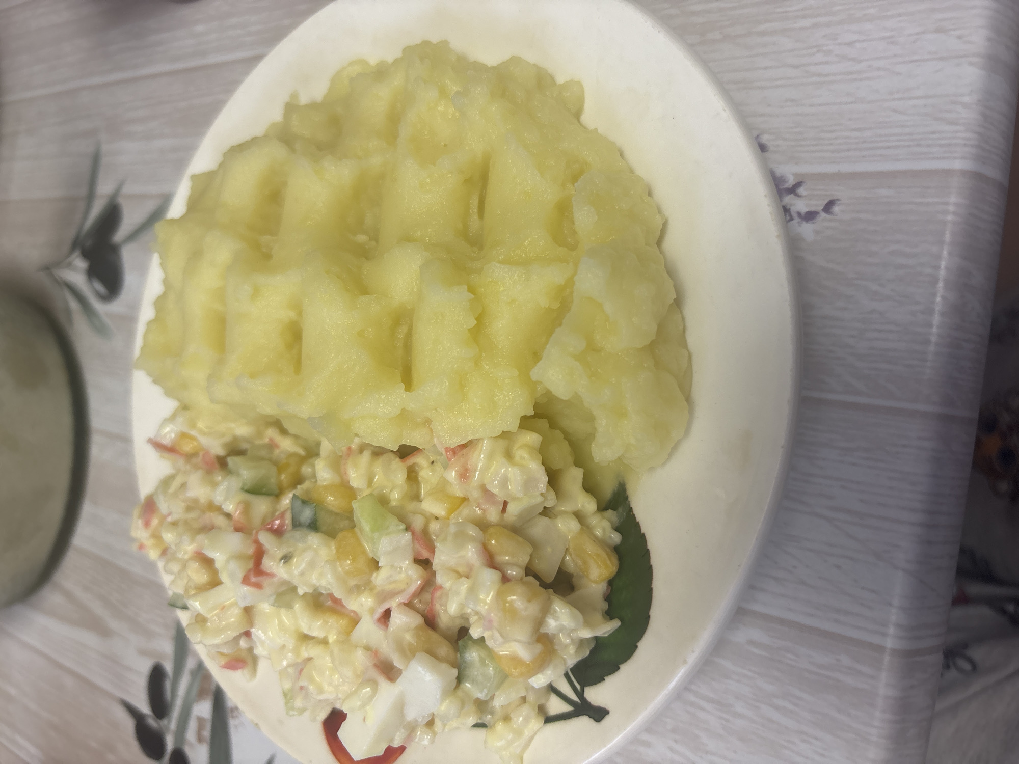 A close-up, overhead shot shows a white plate with a meal on it, likely at a table. The plate contains two distinct food items. The larger portion is a mound of pale yellow mashed potatoes, textured with visible indentations, possibly from a fork or masher. Next to the potatoes is a salad made with a creamy white dressing. The salad appears to contain finely diced ingredients, including what looks like crab sticks (pinkish-red strips), chopped hard-boiled eggs (white cubes), corn kernels (yellow), and chopped cucumber (green).
The plate is positioned on a surface that seems to be a table with a light-colored, wood-grain pattern. The pattern has subtle grey lines and some faint floral or decorative elements in a muted purple color. A hint of another object, possibly a glass or bowl, is visible to the left of the plate. The overall lighting suggests an indoor setting, with no strong indications of time of day or weather. There is no visible text in the image. The meal appears to be a home-cooked dish, ready to be eaten. The setting is likely a kitchen or dining area in Kazan, Russia.