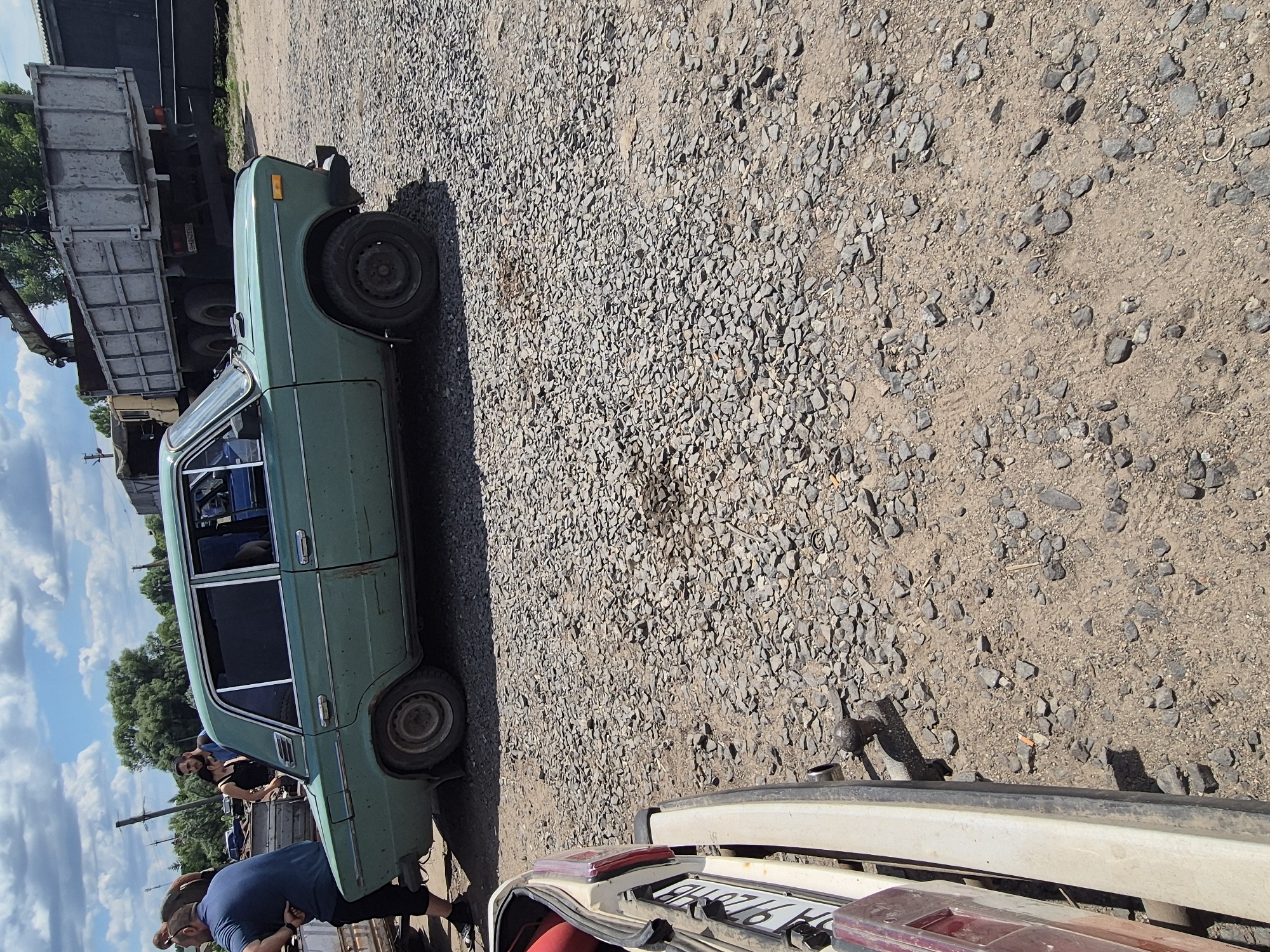 A green, four-door sedan, appearing to be a Lada VAZ-2101, is parked on a gravel lot. The car has visible rust and damage, particularly around the rear quarter panel. In the background, a large, utilitarian truck with a raised bed is visible, along with trees and utility poles. Three individuals are present, two men and a woman, near the left side of the sedan. One man in a blue t-shirt and black pants stands with his back to the camera. Another man, partially obscured, stands behind him. A woman with dark hair, wearing a black tank top and jeans, is also present. The scene appears to be outdoors in what could be a junkyard or a service area. The setting is consistent with the industrial outskirts of a city like Pidhaitsi, Ukraine, given the presence of such vehicles and the overall rugged environment.