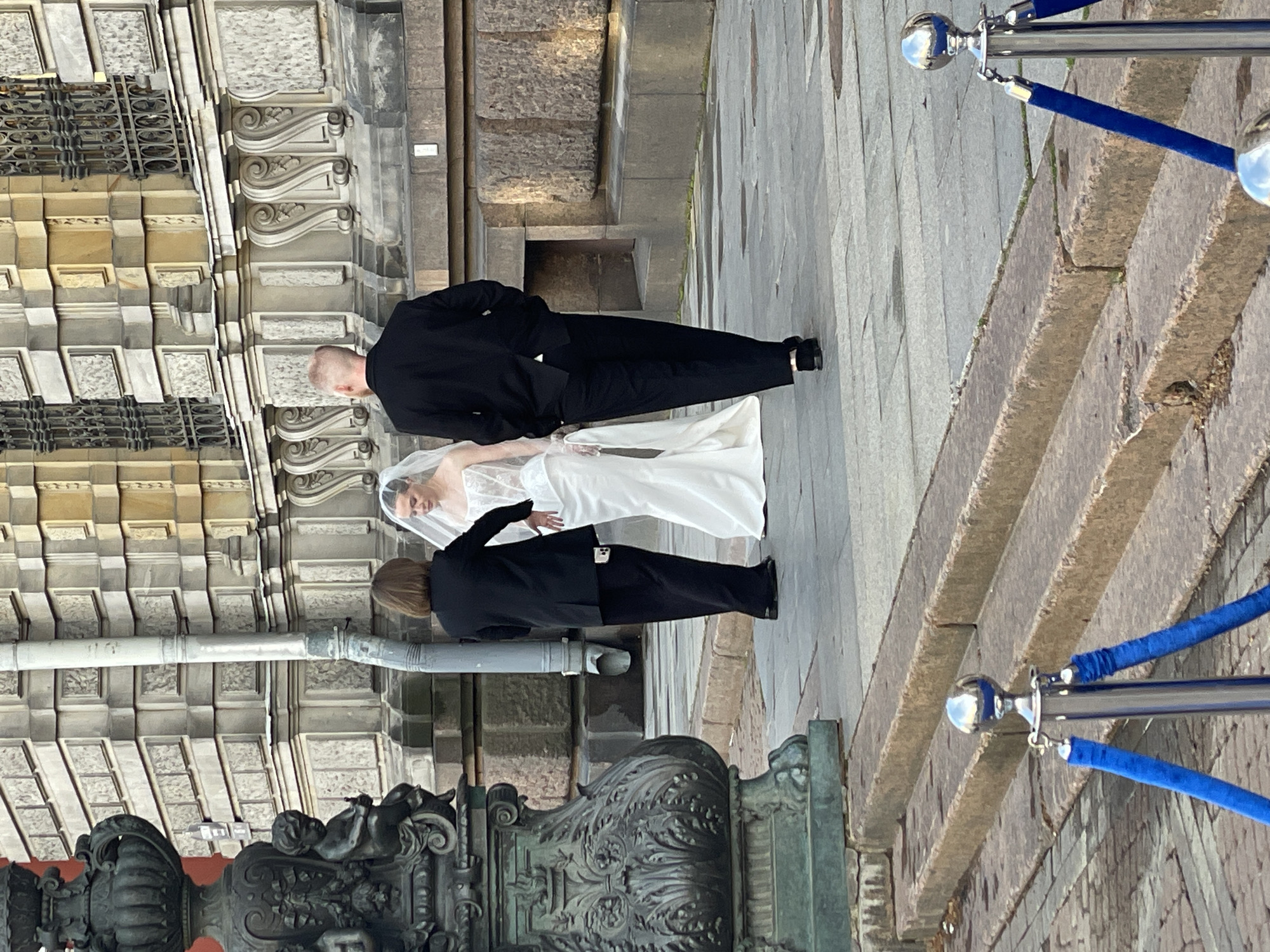 This sideways image depicts a wedding scene outdoors in an urban setting, likely Saint Petersburg, Russia. Three figures are visible on a paved stone surface, standing before a grand, elaborately decorated building.
In the foreground, a man with very short, light-colored hair is dressed in a black suit, standing a little ahead of the others. He is presumed to be the groom. Behind him, a woman in a flowing white wedding gown and a veil stands, looking towards the man and the building. She is presumably the bride. To the bride's side and slightly behind her, a third individual in a black suit with shoulder-length brown hair is present.
The backdrop is a magnificent, light-colored stone building exhibiting classical or baroque architectural features, including ornate pilasters, intricate carvings, decorative cornices, and detailed window grilles. To the left foreground (when rotated correctly), a section of an ornate dark metal structure, possibly a lamp post base or a decorative railing featuring cherub-like figures and elaborate scrollwork, adds to the historical character of the location.
To the right of the figures, a series of stone steps ascends, bordered by blue velvet queue ropes attached to polished chrome stanchions, suggesting a designated pathway or a special event area. The ground consists of large stone pavers, which appear slightly damp in places. The lighting is diffused, indicating it is daytime, possibly an overcast day. No discernible text is visible on any of the elements in the image. The overall impression is a formal and celebratory event set against a backdrop of impressive historical architecture.