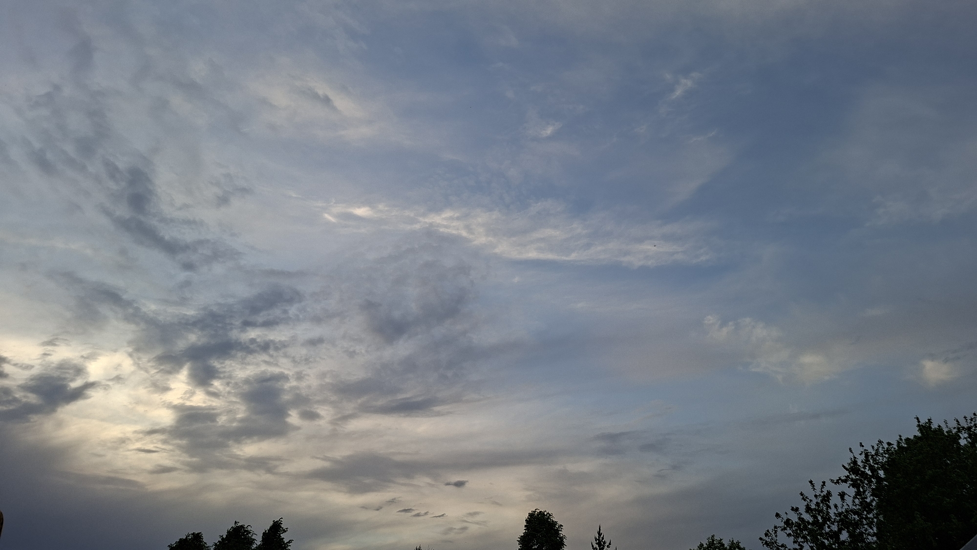 The image shows a cloudy sky. The clouds are various shades of gray and white, with some blue sky visible in between. The lower portion of the image shows trees. The location is presumably Большой Букор, Russia, as specified, but no elements in the image specifically confirm that.