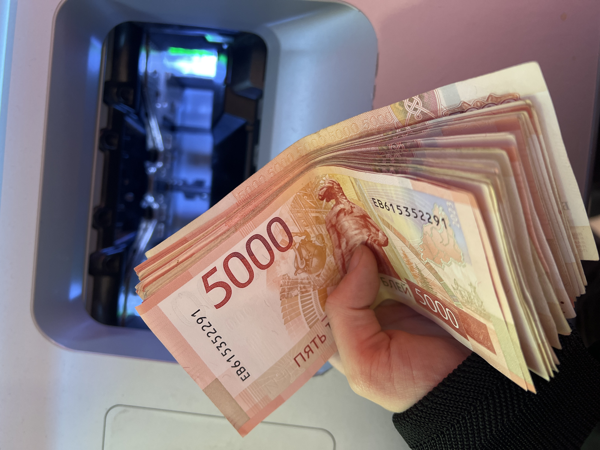 A hand holds a stack of 5000 Russian Ruble banknotes outside of an ATM. The ATM's interior is visible with metallic and blue components. The setting may be Khimki, Russia, based on the currency type.