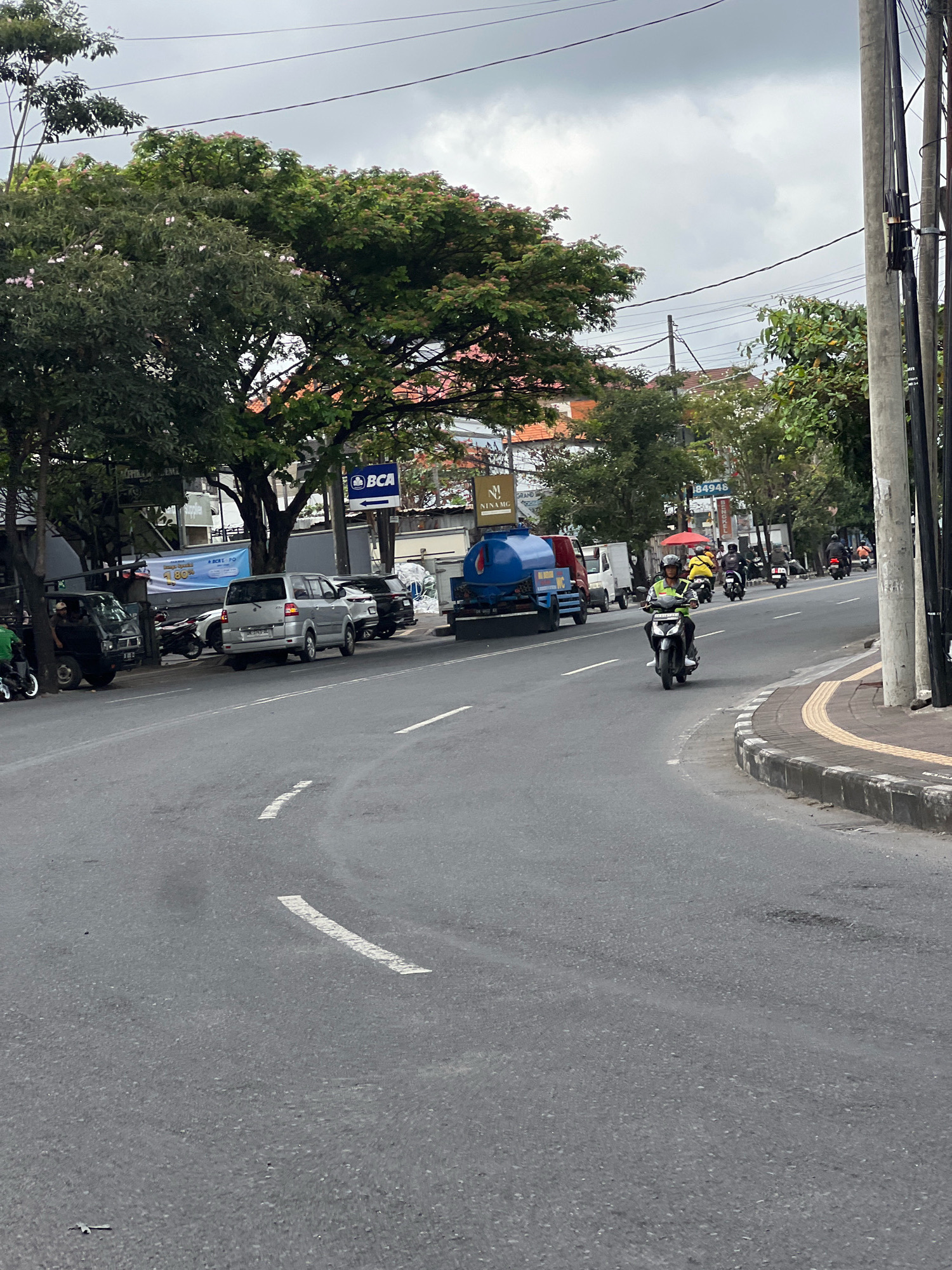 The image depicts a street scene in Denpasar, Indonesia, with traffic and pedestrians. A person is riding a motorcycle in the foreground, wearing a yellow jacket and a helmet. Several other motorcycles and cars are visible on the road and parked along the side. Trees line the street, and buildings with tiled roofs are in the background. A blue water tank truck is parked on the side of the road. The sky is overcast, suggesting a cloudy day. A sign for "BCA" (Bank Central Asia) is visible. The road is paved with asphalt and has white lane markings. The sidewalk is lined with concrete and has a yellow tactile paving.