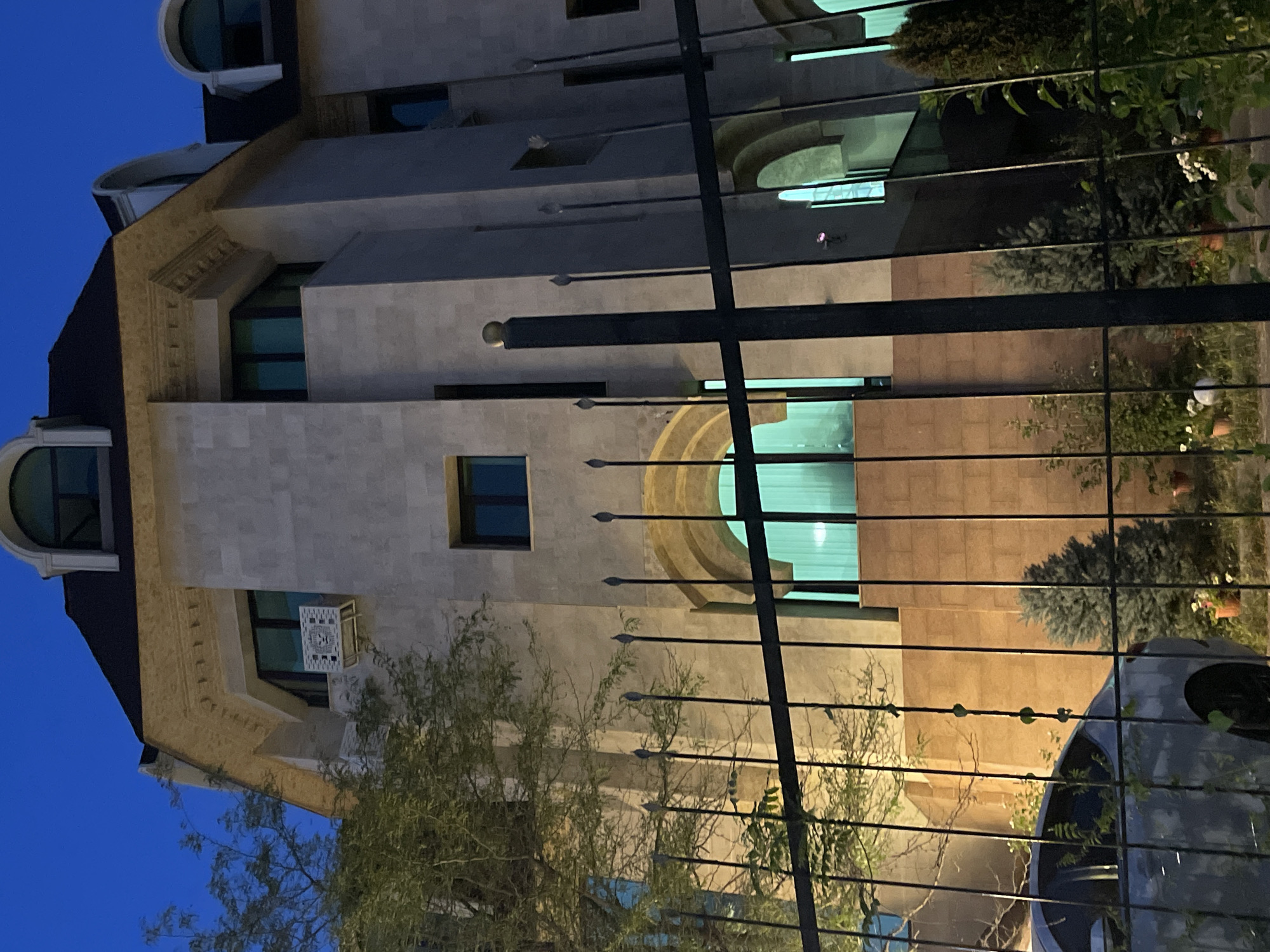 The image depicts a stone-clad building at dusk in Almaty, Kazakhstan. A black metal fence runs across the foreground, partially obscuring the view. Through the fence, a silver car is visible on the left, and several small evergreen trees and potted plants are scattered in the yard. The building itself features arched windows with teal-colored curtains and a dark pitched roof with dormer windows. An air conditioning unit is mounted on the exterior wall. The sky is a deep blue, indicating the transition from day to night. The overall scene suggests a residential or commercial property in a city setting.