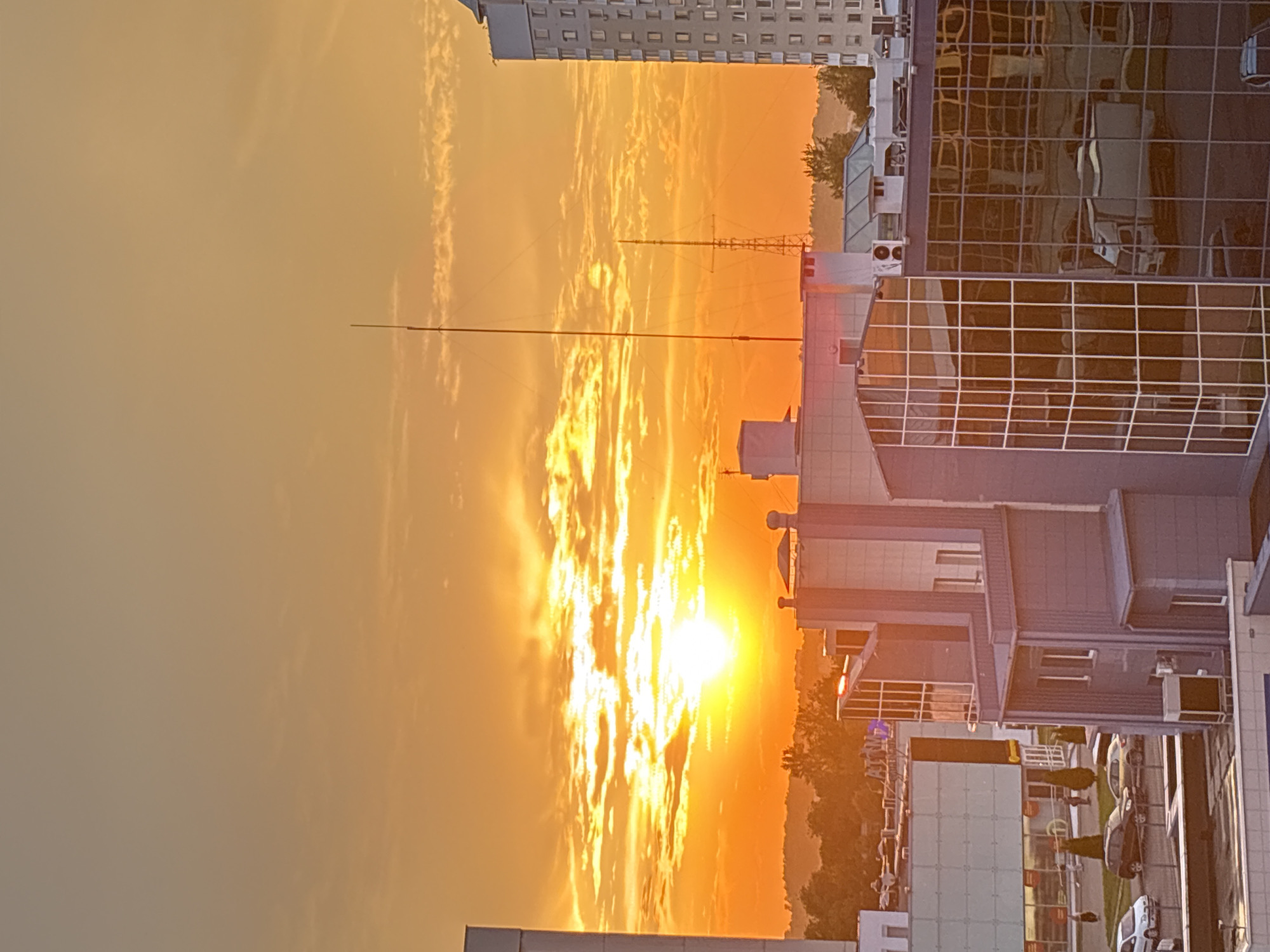 The image depicts a sunset over the city of Vitsebsk, Belarus. The sky is filled with vibrant orange and yellow hues, with the sun casting a bright glow as it descends. Several buildings are visible in the foreground, including modern commercial structures with reflective glass facades and older, multi-story buildings. One prominent building on the right is tall and gray with numerous windows. In the middle ground, a medium-sized building with white and gray sections features a large glass-paneled section. Smaller buildings and structures, along with trees, are visible in the distance, leading to a treeline on the horizon. A few cars are parked in a lot in front of one of the buildings. Two tall antennas or communication towers are also present in the scene, silhouetted against the sky. The overall impression is of an urban landscape at dusk, bathed in the warm light of the setting sun.