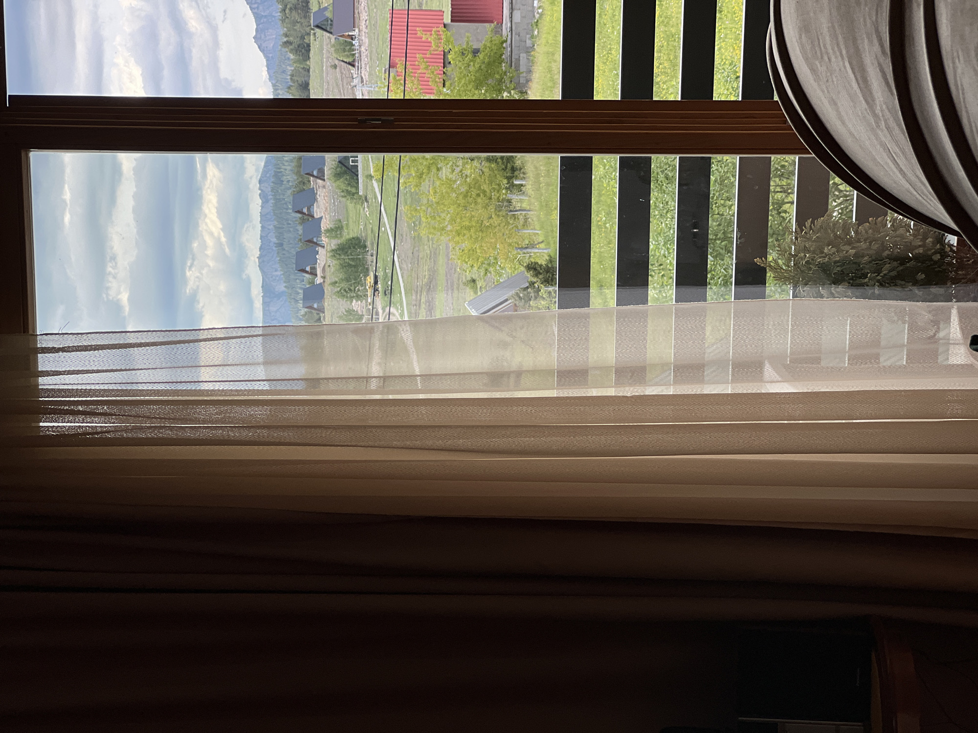The image presents an interior view looking outwards through a large window, framed by drawn curtains. On the left, a heavy, dark brown curtain covers the wall, partially obscuring a dark electronic device on a wooden surface in the lower-left corner. A translucent, light beige sheer curtain with a subtle herringbone pattern is drawn across the right portion of the window, filtering the exterior light. The window itself has a dark brown wooden frame, vertically segmenting the view.
Immediately outside the window and within the lower frame is a balcony railing composed of multiple horizontal dark bars. Below this railing, a small potted conifer is visible on the balcony, alongside green ground cover.
The exterior scene reveals a rural or semi-rural landscape under an overcast sky with numerous white and grey clouds. In the mid-ground, a gently sloping grassy field is interspersed with trees, some featuring bright green deciduous foliage, others darker conifers. Several identical A-frame houses with dark roofs and light walls are clustered on the slope. A dirt or gravel path winds through the field, and a yellow excavator is visible near the center-left. Overhead wires traverse the landscape. To the right, a building with a bright red roof and concrete-like base is partially visible, next to a smaller red structure. In the distant background, a range of mountains, heavily forested with conifers and exhibiting rocky outcrops, extends across the horizon. No individuals or animals are present in the scene.