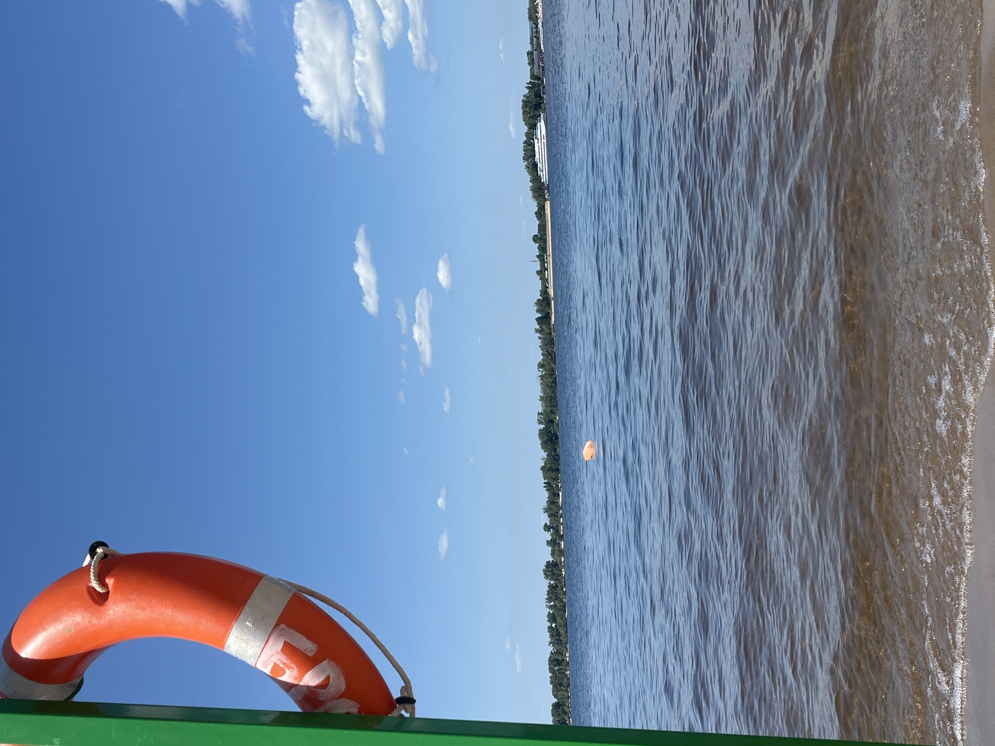 A life preserver hangs from a green pole on the left side of the frame, against a bright blue sky with scattered white clouds. The life preserver is orange with white reflective bands and rope. In the distance, across a calm body of water, a treeline runs along the horizon, suggesting a shoreline. A large white boat is visible on the water near the far shore. There is a small orange buoy floating in the water closer to the foreground. The water's surface is rippled, and the bottom is visible in the shallows, indicating a sandy or pebbly shore. The scene appears to be a waterfront area, possibly a beach or pier, on a sunny day. The specific location in Novokuybyshevsk, Russia cannot be determined from the image alone.